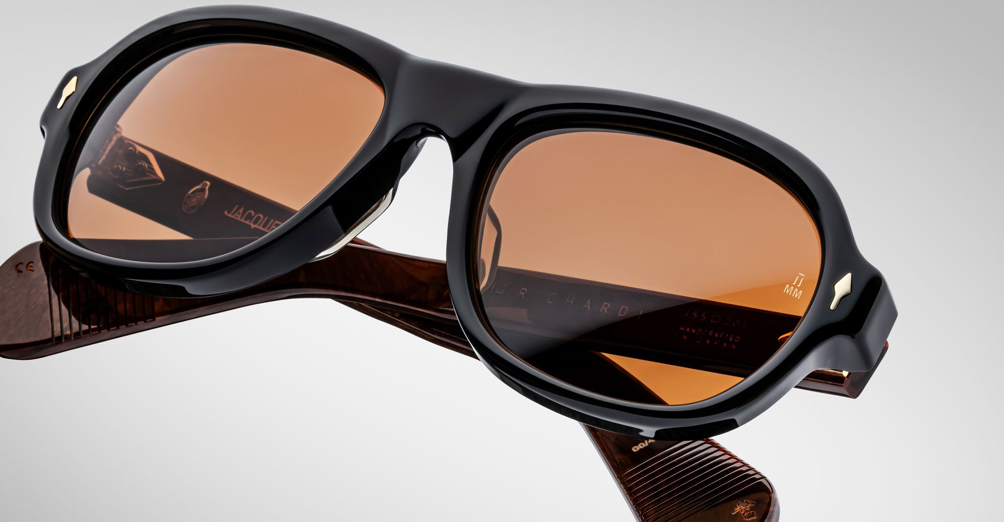 Close-up of Jacques Marie Mage Richard sunglasses with thick, glossy black frames and brown-tinted lenses, resting on their side. The visible arms feature engraved details and text on the inner side.