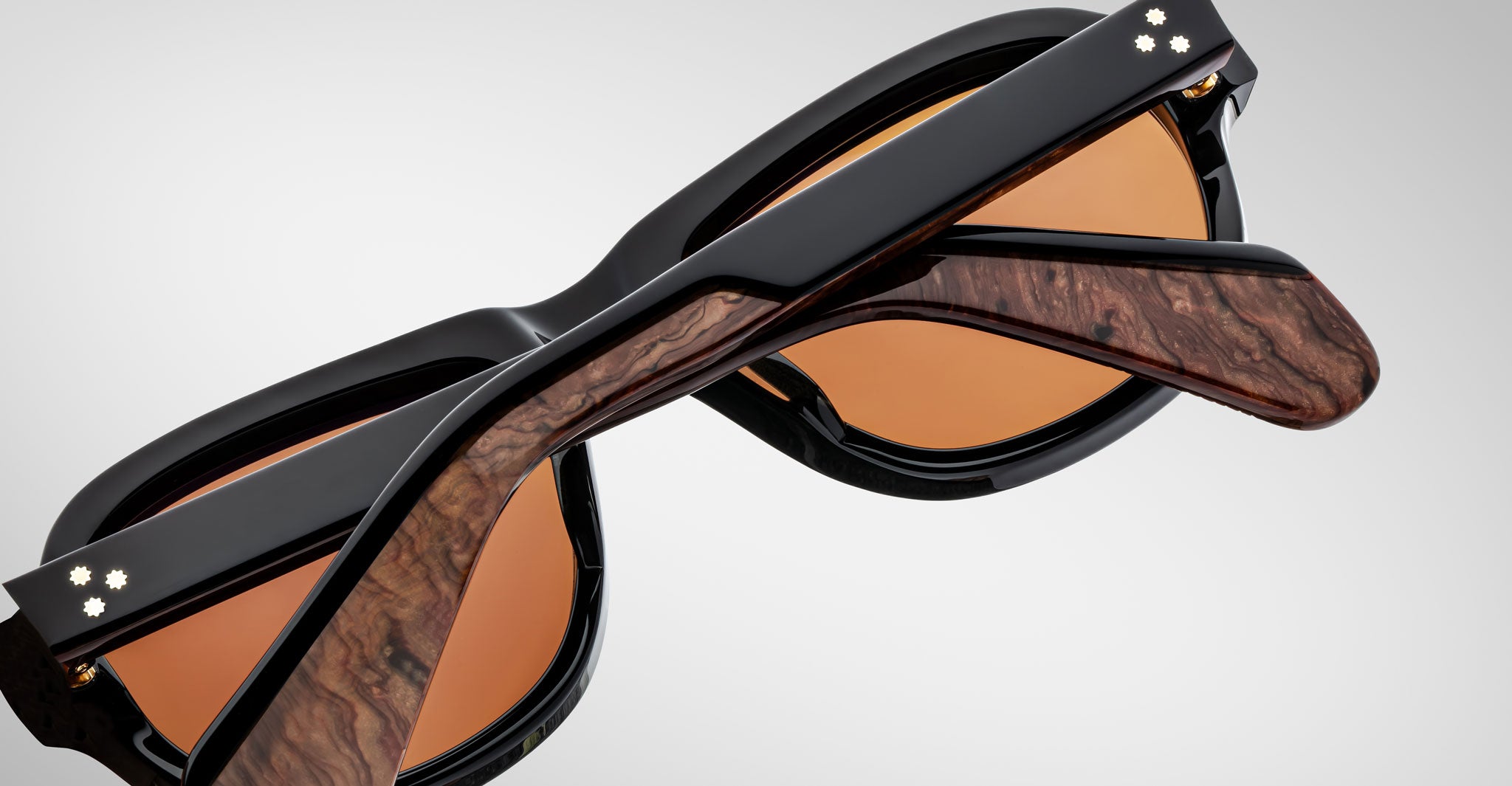 A close-up of two Jacques Marie Mage Richard sunglasses with brown-tinted lenses, black frames, and wood-textured temple accents, shown overlapping on a light gray background.