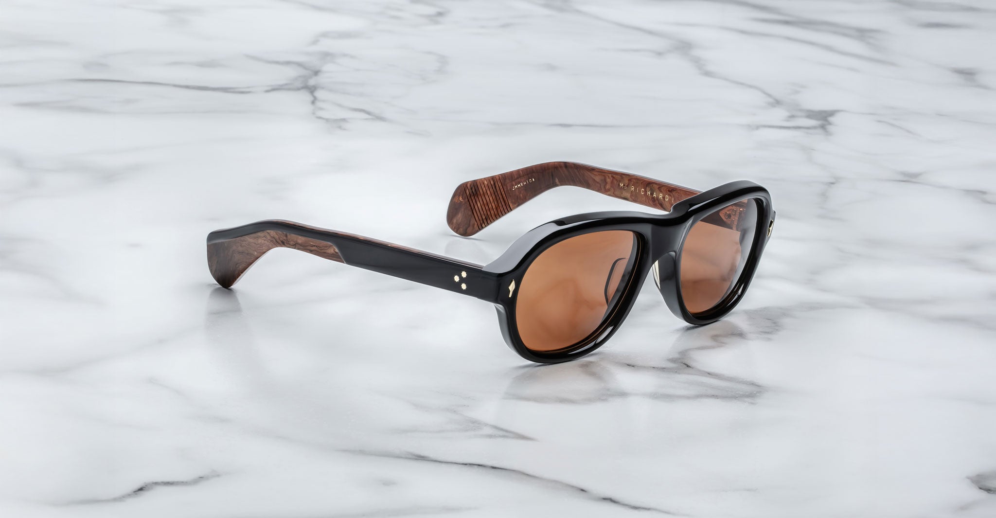 The Jacques Marie Mage Richard sunglasses, featuring black frames, brown-tinted lenses, and wood-textured arm interiors, rest on a white marble surface.