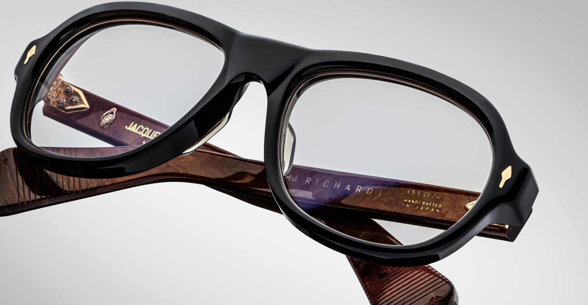 A close-up of Jacques Marie Mage Richard eyeglass frames in black with thick rims, clear lenses, and gold temple details, set against a plain light gray background.