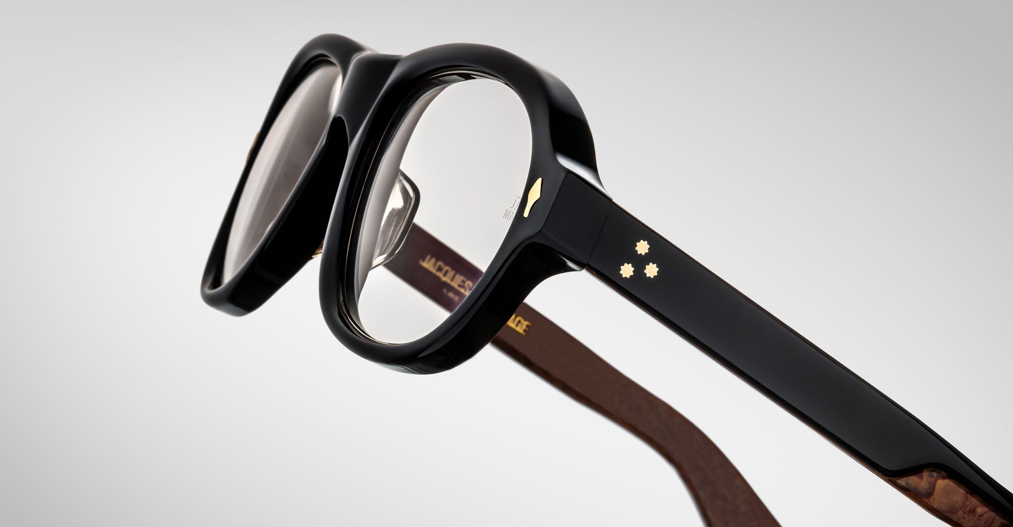 Close-up of Jacques Marie Mage Richard thick black eyeglass frames with clear lenses. The temple arm displays two small gold star-shaped details and a gold chevron, with interior text visible. Background is light gray.