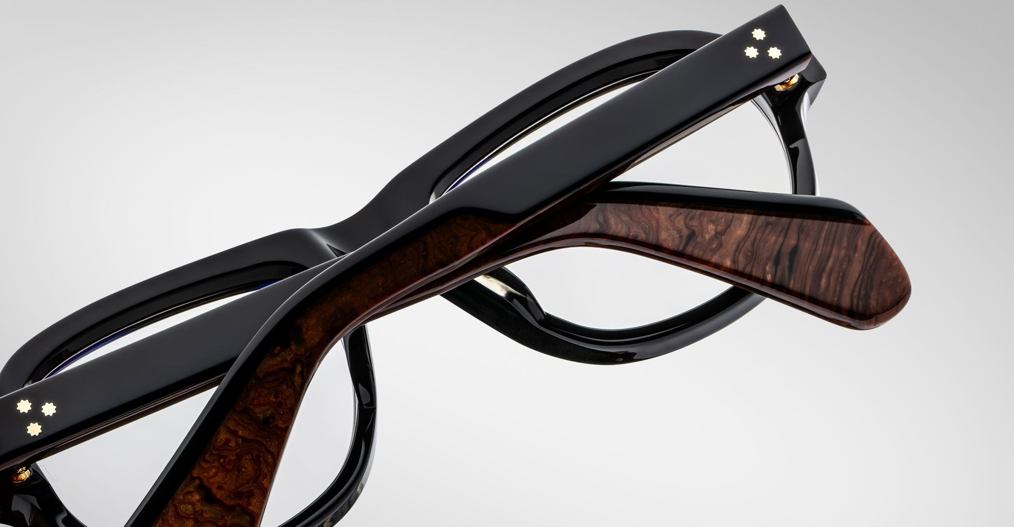 A close-up of two Jacques Marie Mage Richard eyeglasses with black frames—one featuring wood-textured arms and both accented by small decorative dots on the hinges. The background is a plain light gray.