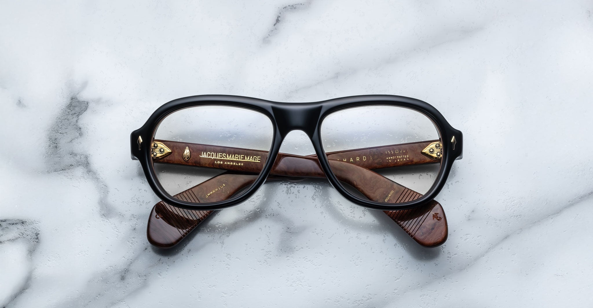 A pair of Jacques Marie Mage Richard eyeglasses with black rims and brown temples is placed upside down on a white marble surface, showing gold text and markings inside the temples.
