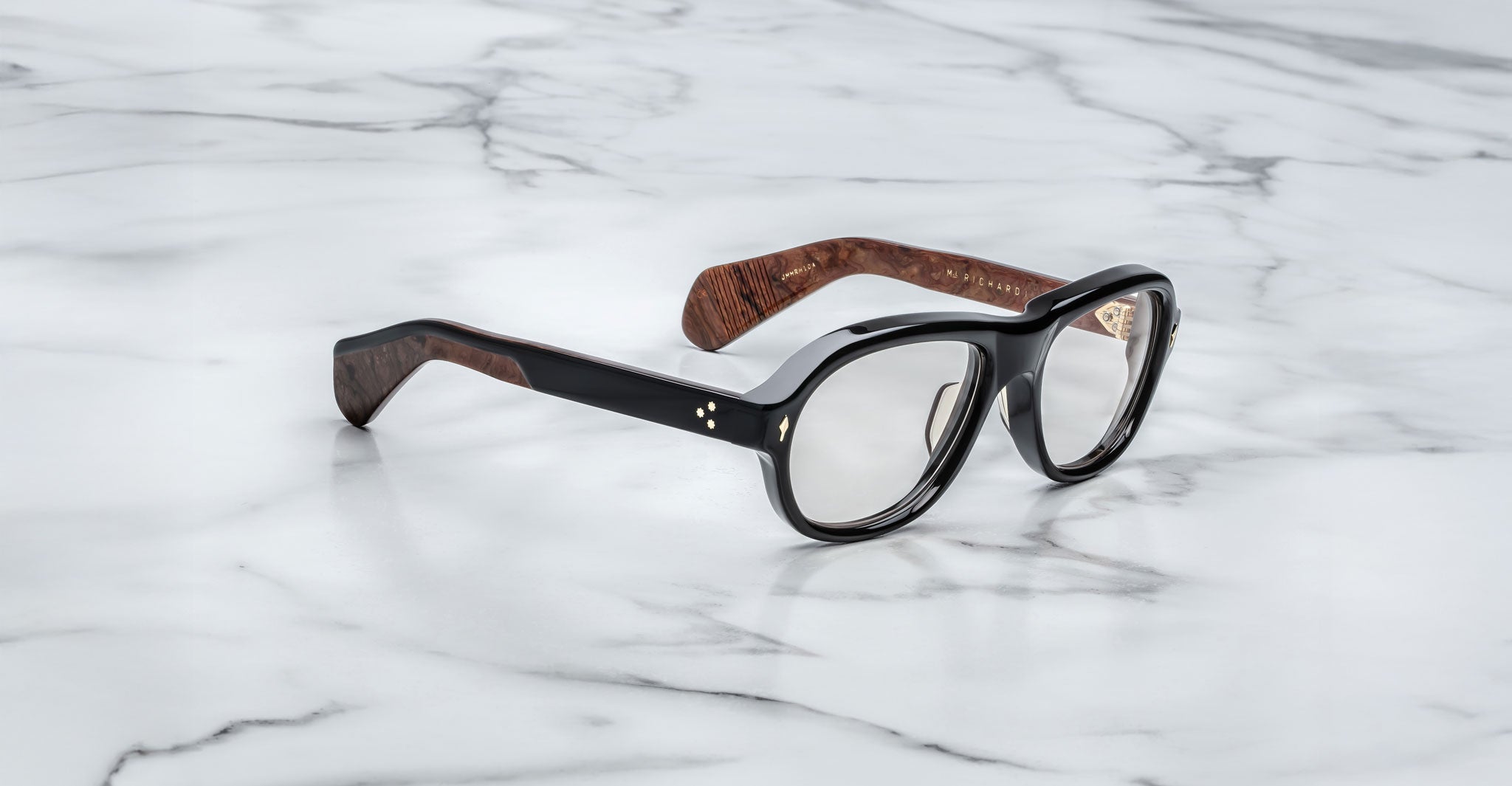 The Jacques Marie Mage Richard eyeglasses, featuring thick black and brown frames, rest on a white marble surface.
