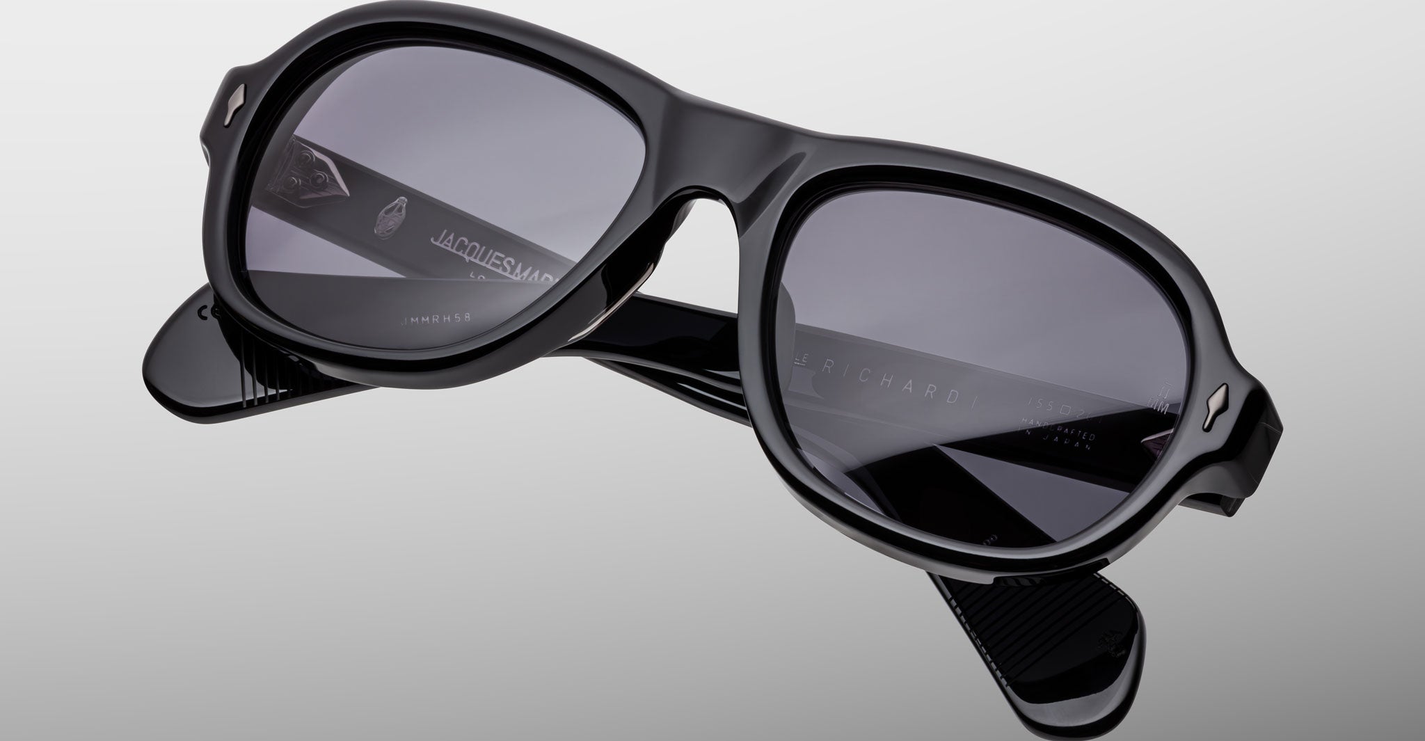 The Jacques Marie Mage Richard sunglasses feature thick, glossy black frames with dark lenses and visible brand markings on the arms, displayed against a light gray background.