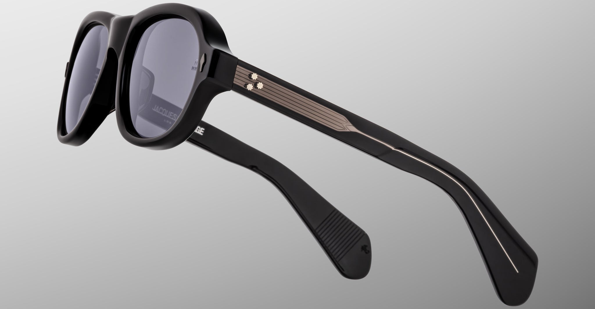 The Jacques Marie Mage Richard sunglasses feature thick black frames, dark lenses, and metallic gold accents with lined temples, displayed on a light gray background.