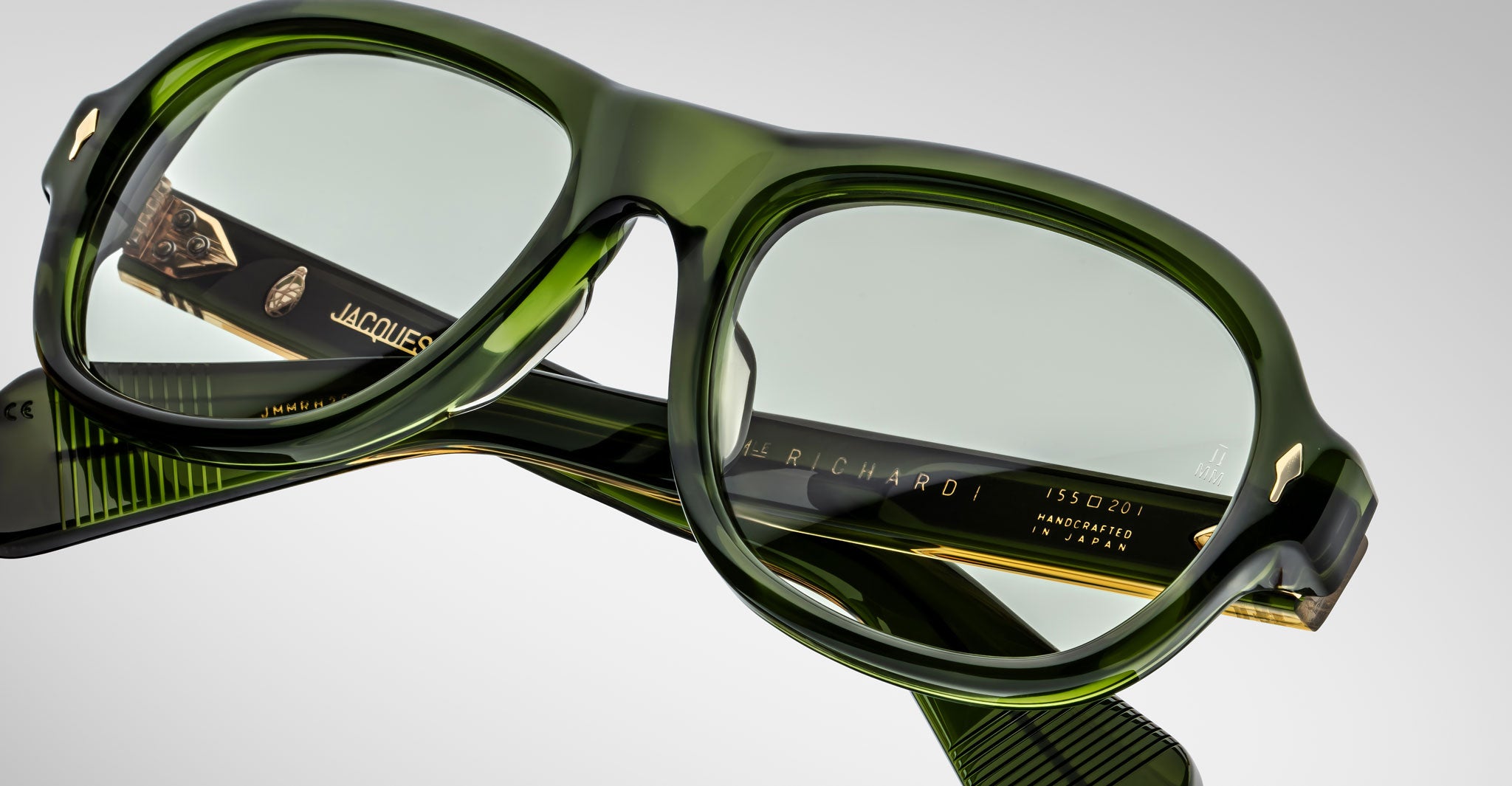 A close-up of green translucent Jacques Marie Mage Richard eyeglasses with clear lenses, gold accents, and visible branding on the arms, including JACQUES, RICHARD, and MADE IN JAPAN.
