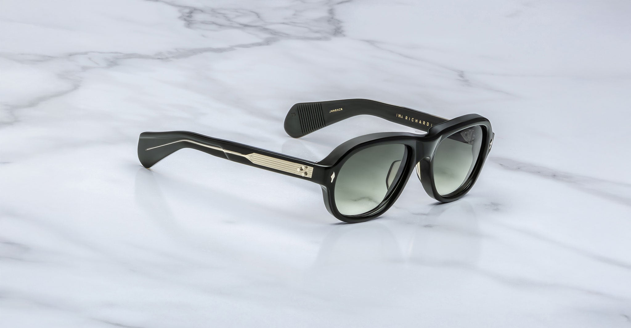A pair of Jacques Marie Mage Richard black sunglasses with dark lenses and gold detailing on the arms rests on a white marble surface.
