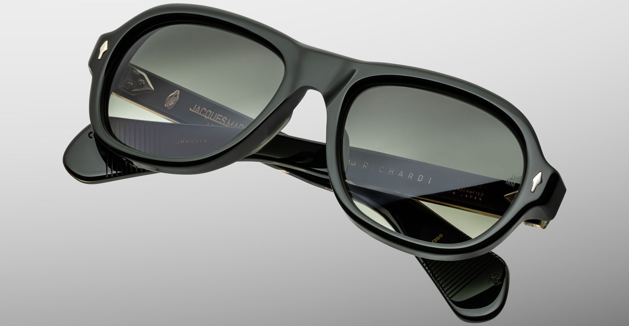 A close-up of Jacques Marie Mage Richard stylish black sunglasses with dark tinted lenses, gold accents, and branded text on the arms, set against a plain light gray background.