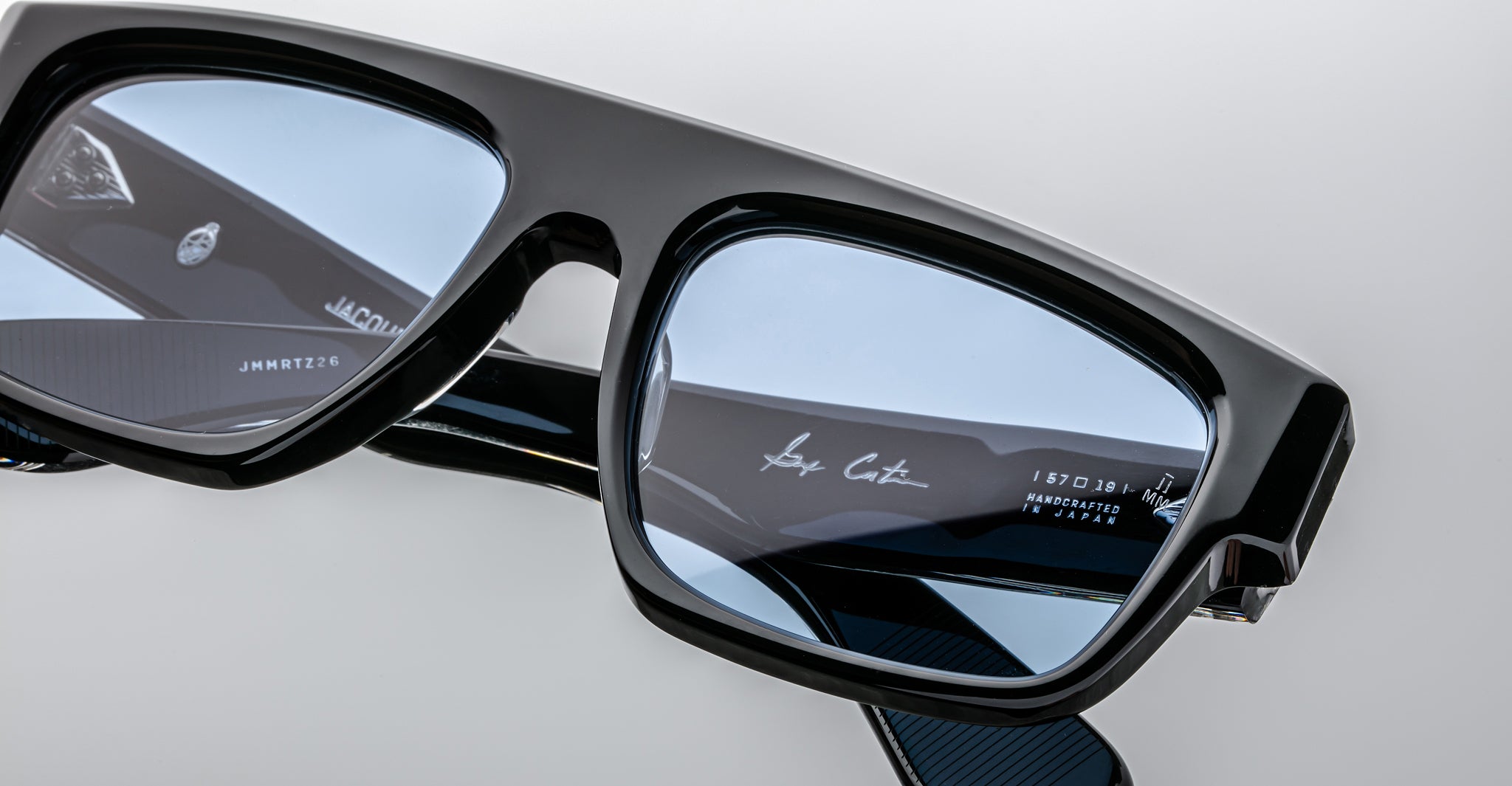 Close-up of Jacques Marie Mage Ritz black rectangular eyeglass frames with blue-tinted lenses, highlighting detailed logos and inscriptions on the arms and lens, set against a plain light background.