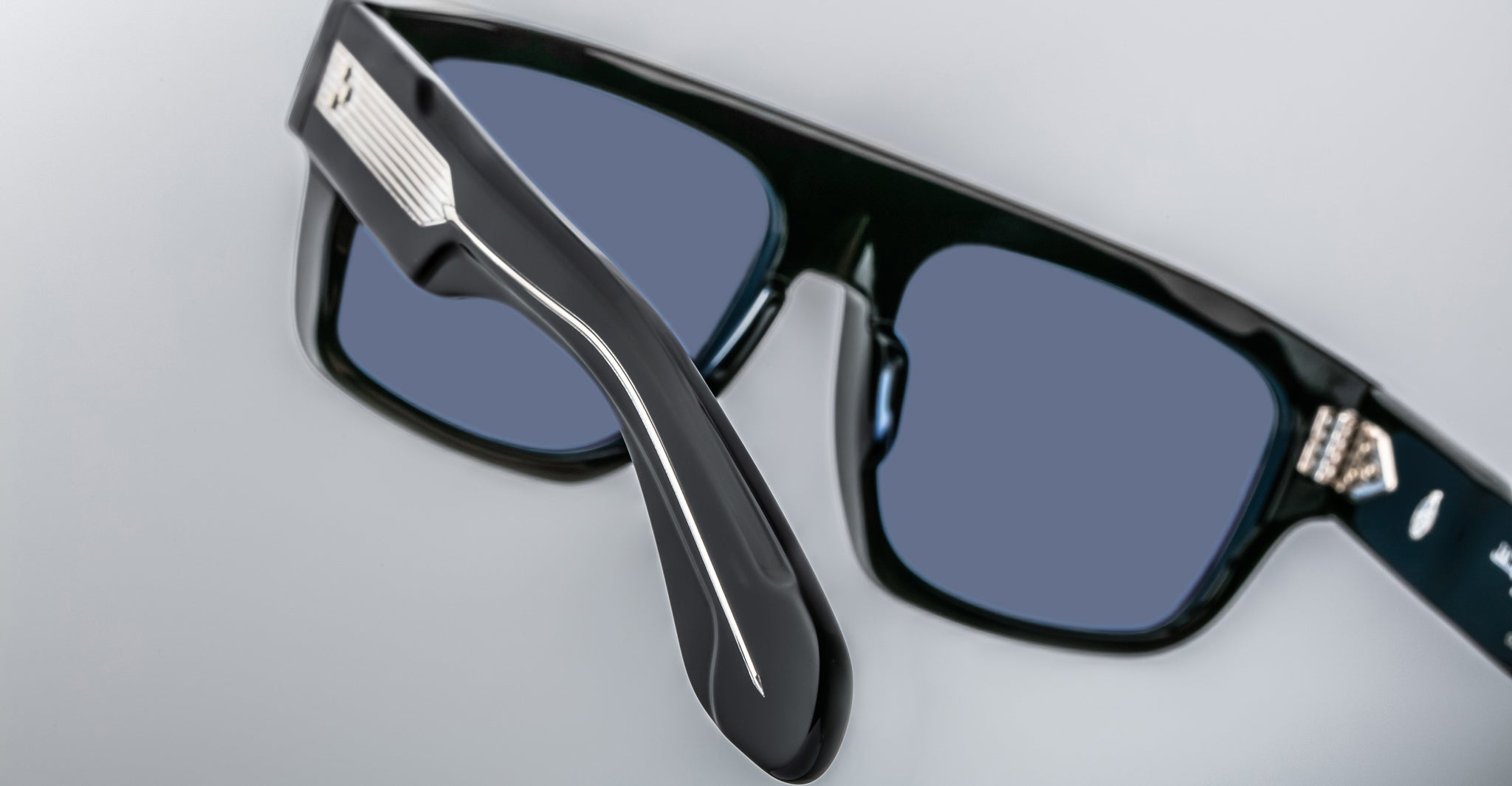 Close-up photo of Jacques Marie Mage Ritz black sunglasses with thick frames, dark blue lenses, and a metallic hinge detail on the arm, set against a plain light background.