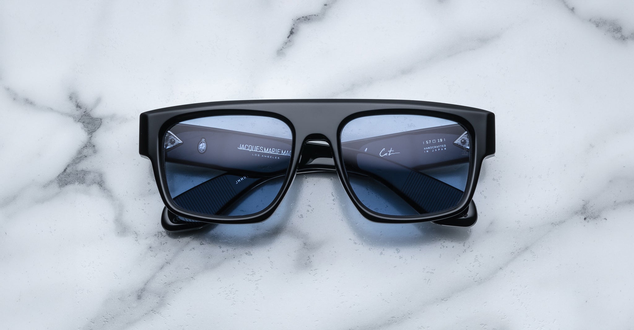 A pair of Jacques Marie Mage Ritz black rectangular sunglasses with thick frames and dark lenses rests on a white marble surface, with reflections visible on the lenses.