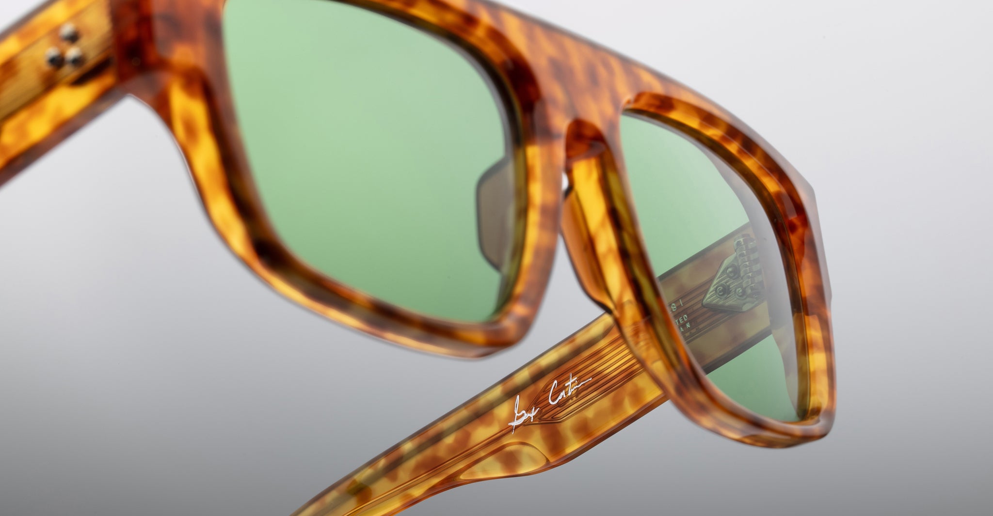 Close-up of Jacques Marie Mage Ritz sunglasses featuring a tortoiseshell frame, green lenses, and a white signature on the temple, set against a neutral light gray background.