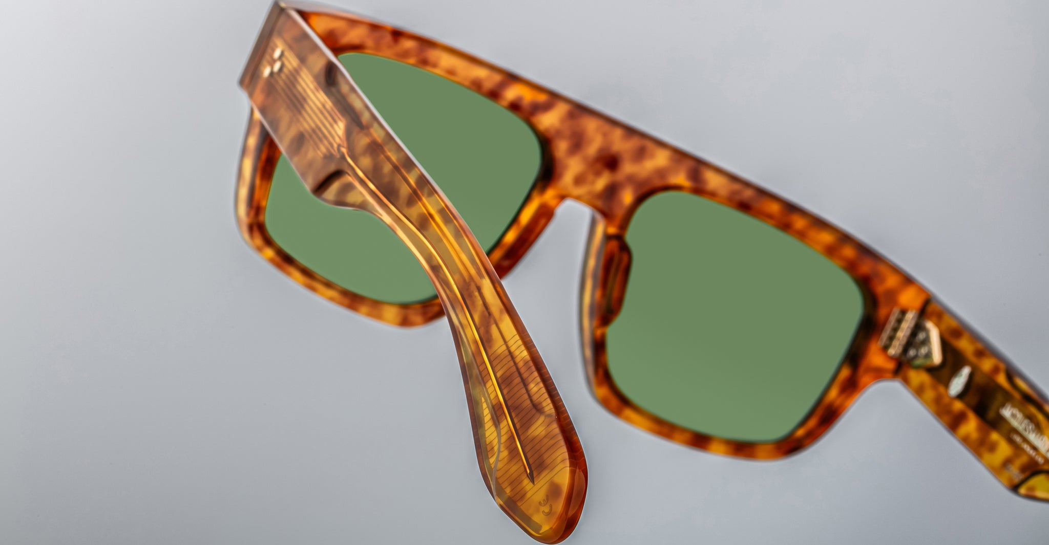 Close-up view of Jacques Marie Mage Ritz tortoiseshell sunglasses with green lenses, one arm extended and the other partially visible, set against a plain light gray background.