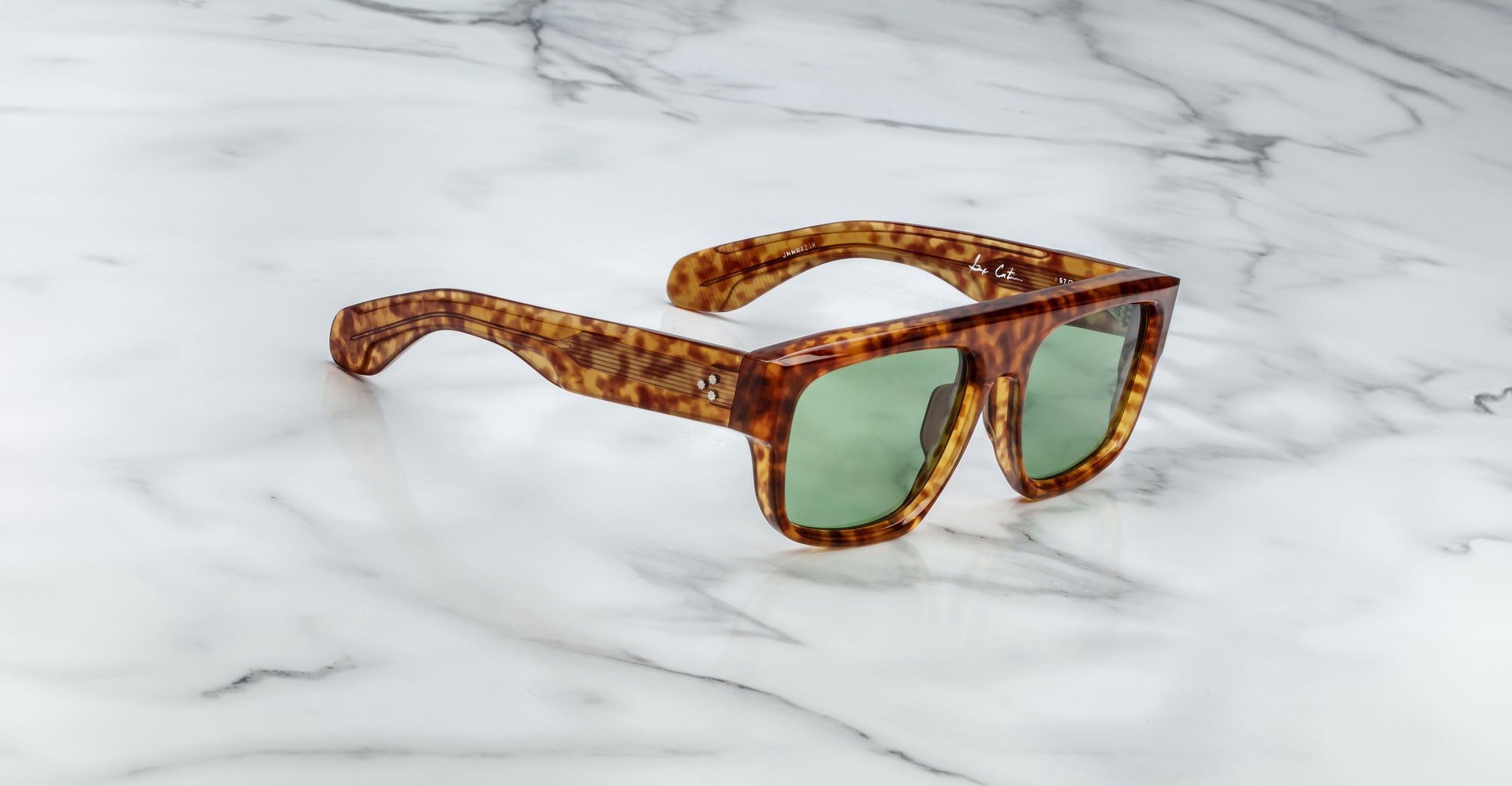 The Jacques Marie Mage Ritz sunglasses, featuring thick brown tortoiseshell frames and green-tinted lenses, rest on a white marble surface with gray veining.