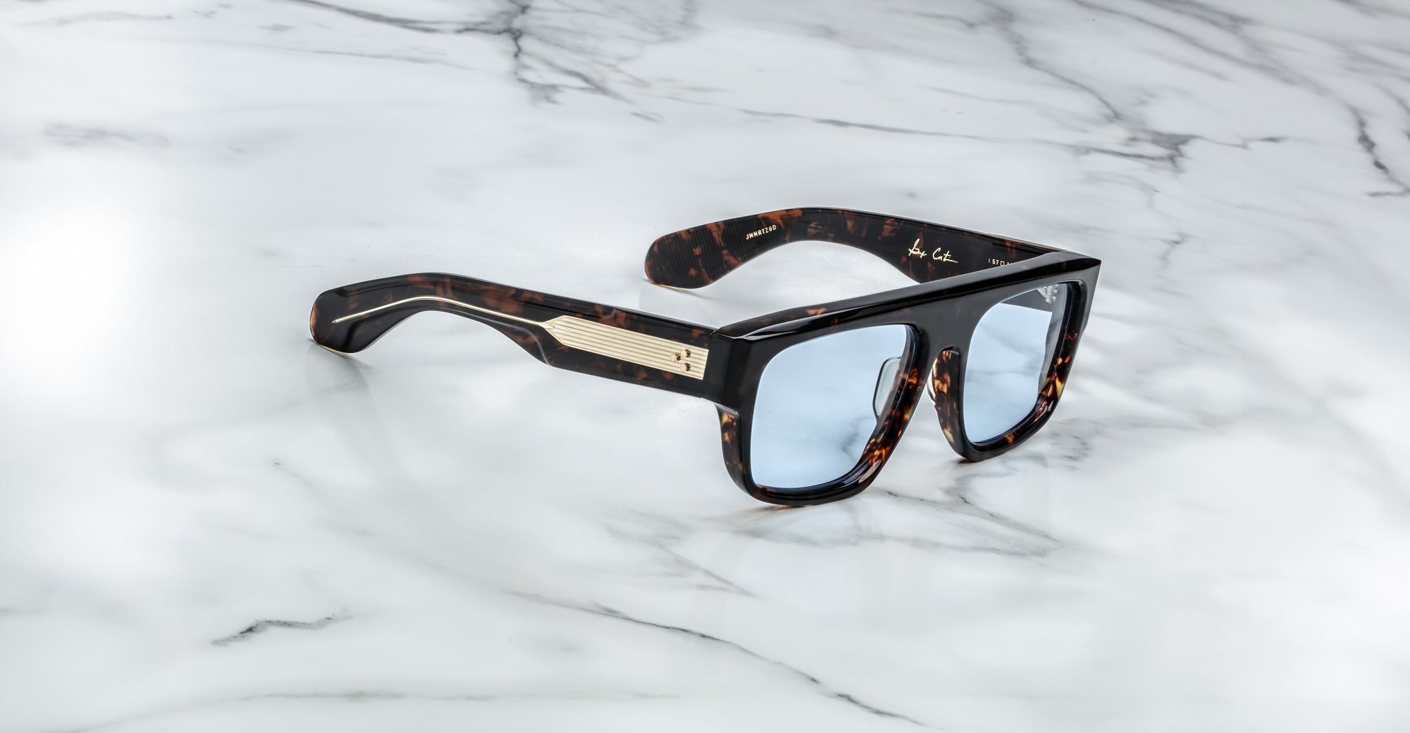 The Jacques Marie Mage Ritz are thick tortoiseshell sunglasses with blue-tinted lenses and gold accents on the temples, displayed on a white marble surface.