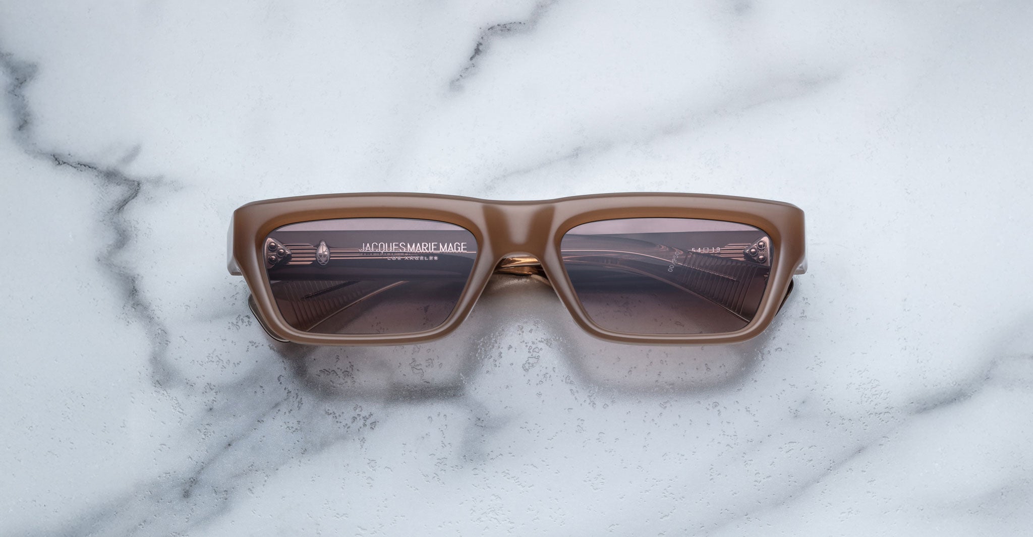 A pair of Jacques Marie Mage Rush brown rectangular sunglasses rests on a white marble surface, their lenses reflecting parts of a building structure with visible lines and text.