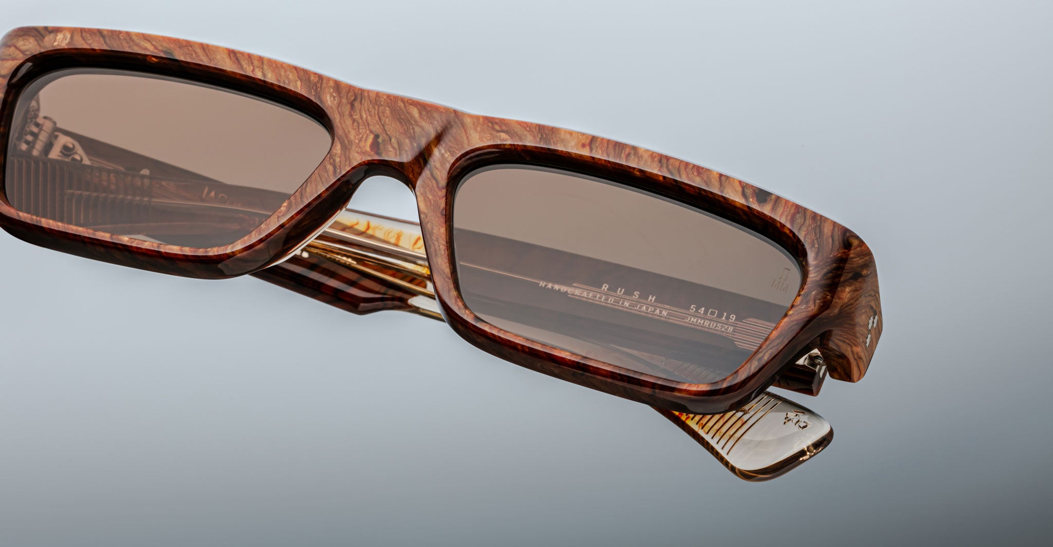 A close-up of Jacques Marie Mage Rush sunglasses featuring thick, brown marbled rectangular frames with grain-like patterns, gold accents, and brown tinted lenses set against a neutral gray background.