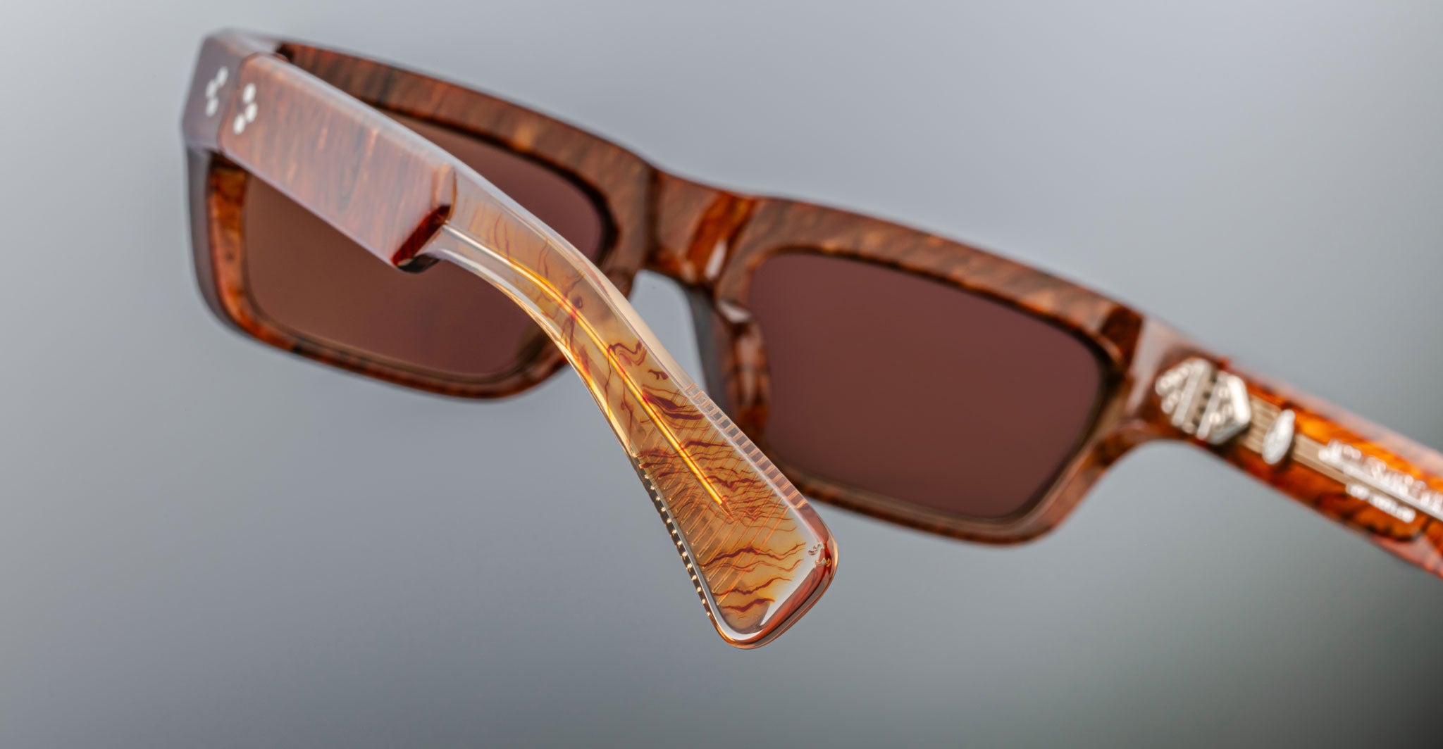 Close-up of Jacques Marie Mage Rush brown rectangular sunglasses with marbled frames and arms. The image highlights the hinge and detailed texture of the arm against a neutral gray background.