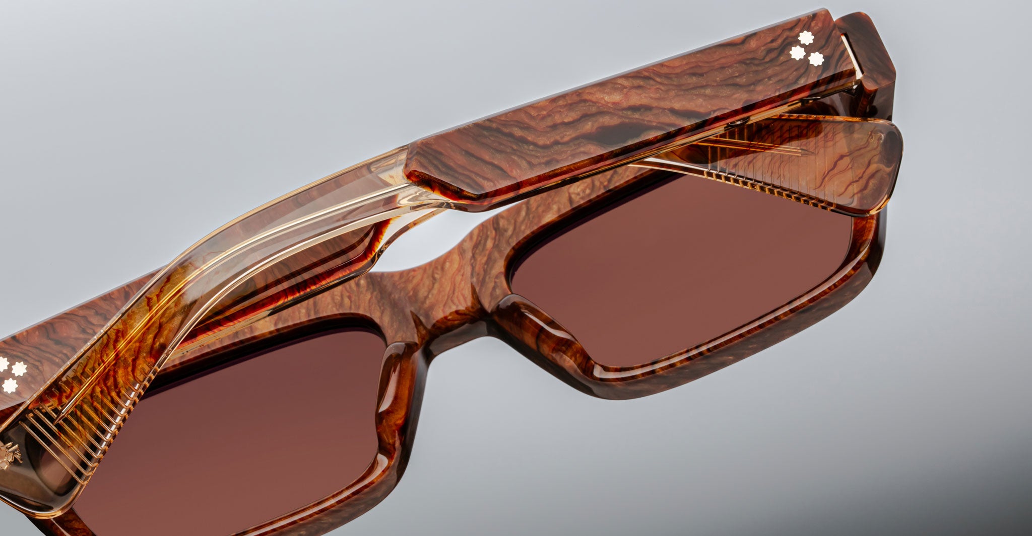 A close-up of the Jacques Marie Mage Rush rectangular sunglasses, featuring brown woodgrain-patterned frames and matching brown-tinted lenses, displayed against a plain gray background.