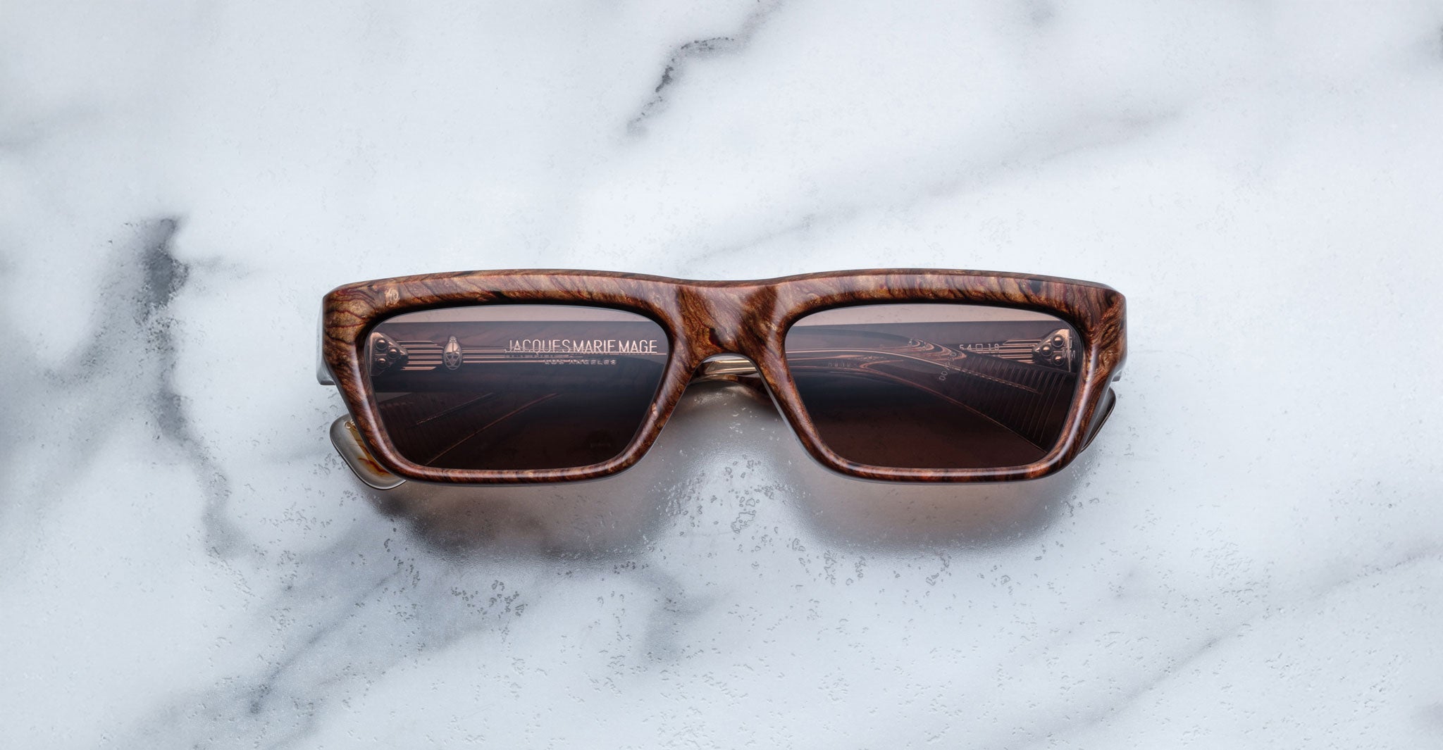 A pair of Jacques Marie Mage Rush rectangular brown sunglasses with dark lenses and a wood-textured frame is placed face-up on a white marble surface. The brand name appears faintly inside the right lens.