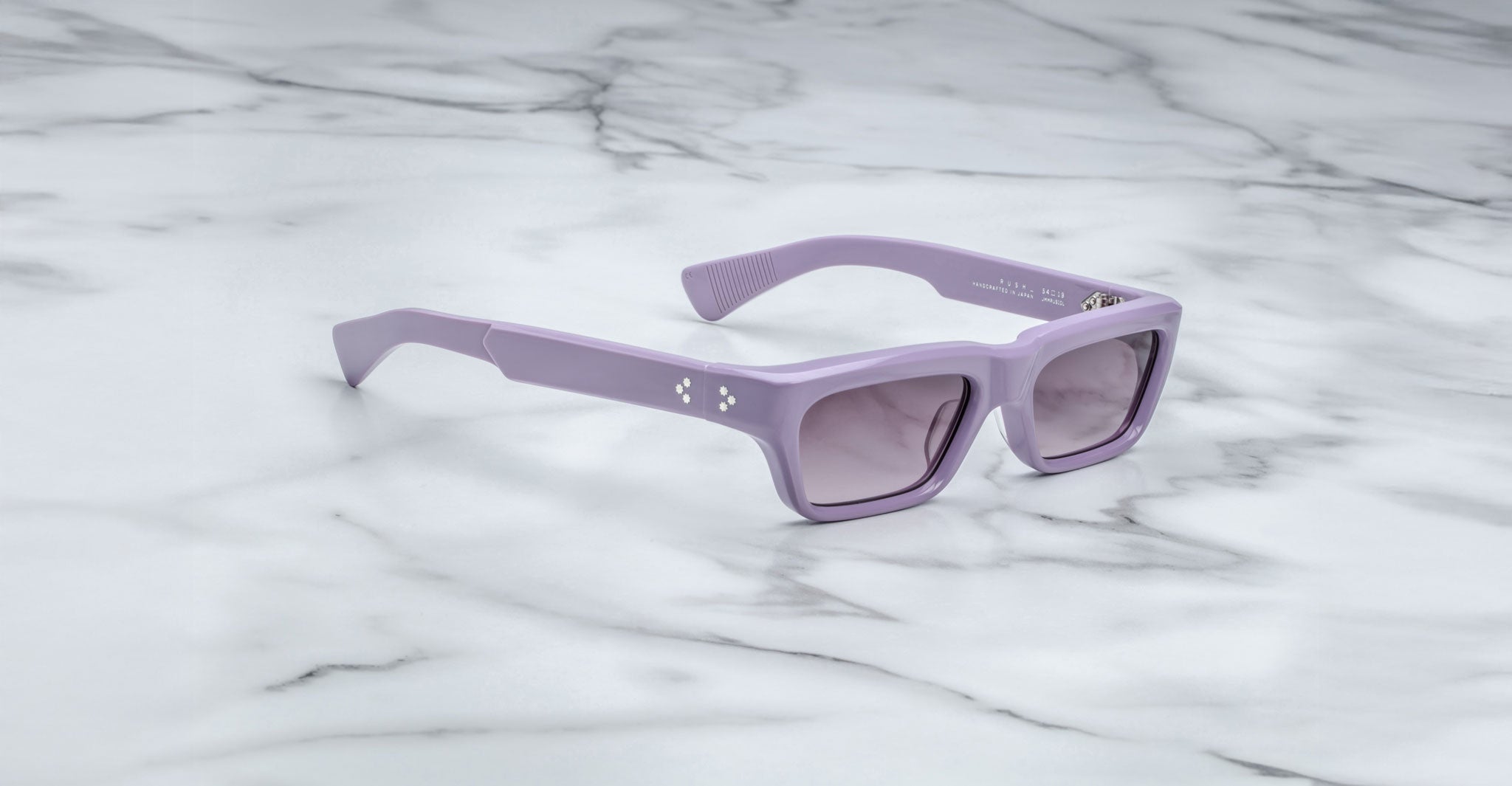 The Jacques Marie Mage Rush sunglasses, featuring rectangular lavender frames with a glossy finish and small white dot pattern on the temple, rest on a white marble surface. The tinted lenses complete the chic look.