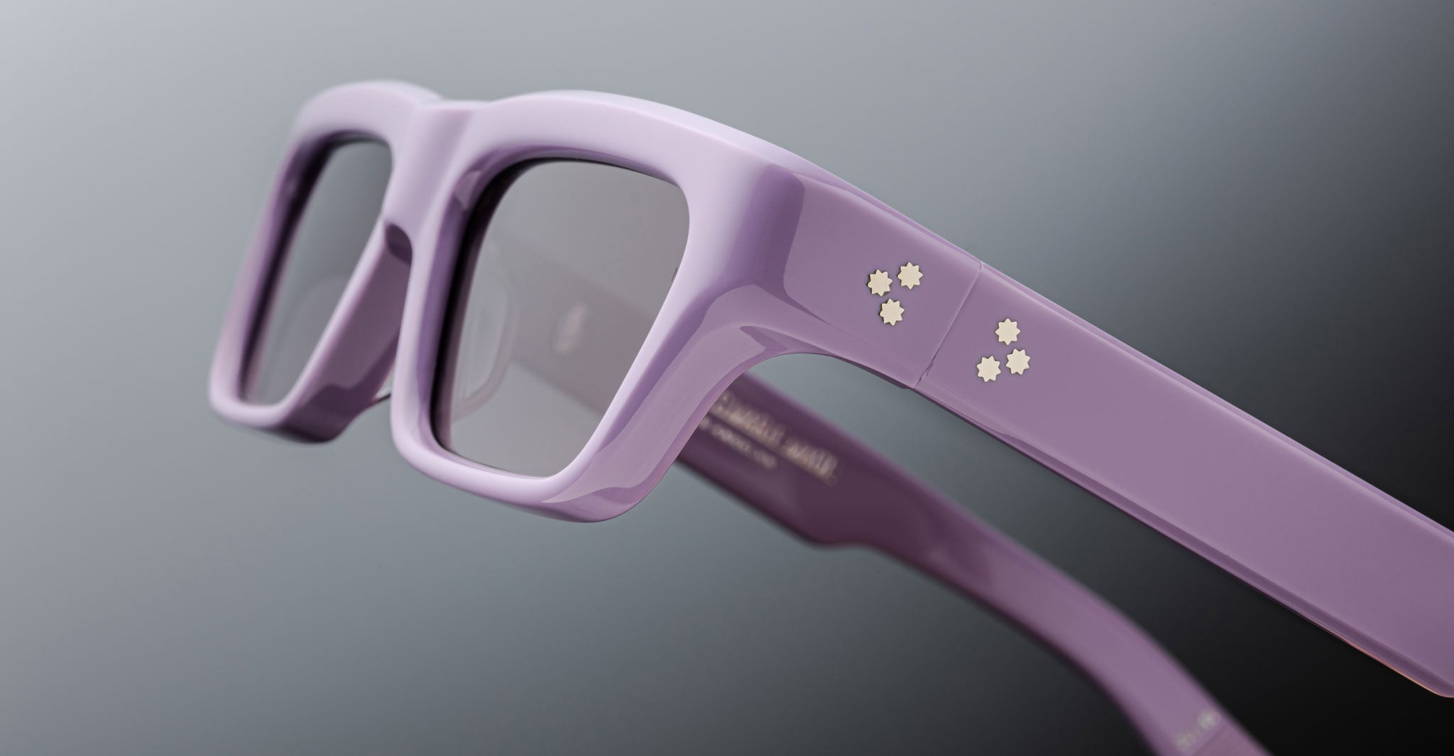 Close-up of Jacques Marie Mage Rush eyeglasses in purple with clear lenses, featuring four white dots on the temple. The background is a soft gray gradient.