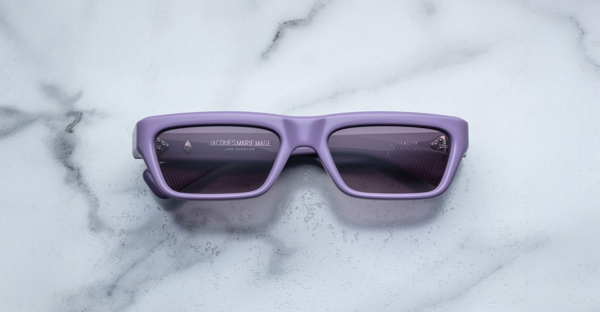 The Jacques Marie Mage Rush sunglasses feature rectangular, thick lavender frames and dark tinted lenses, displayed on a white marble surface.