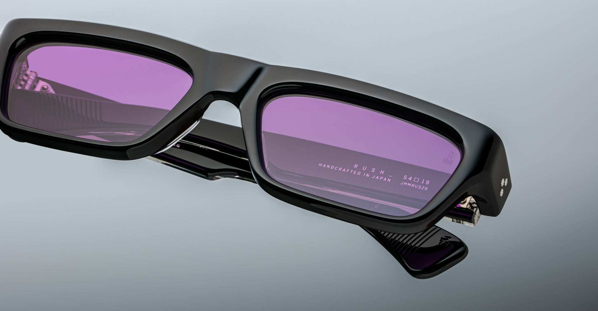 Close-up of Jacques Marie Mage Rush black rectangular eyeglasses with purple-tinted lenses, featuring RUSH and HANDCRAFTED IN JAPAN inscribed on the inner arm against a light gray background.