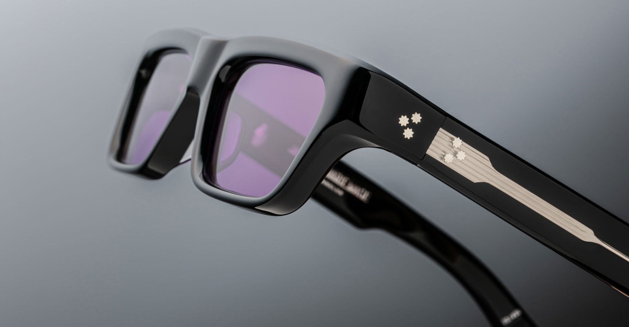 Close-up of Jacques Marie Mage Rush eyeglasses featuring a black rectangular frame, purple lenses, and a metallic detail with three small star shapes on the arm. Set against a plain gradient grey background.