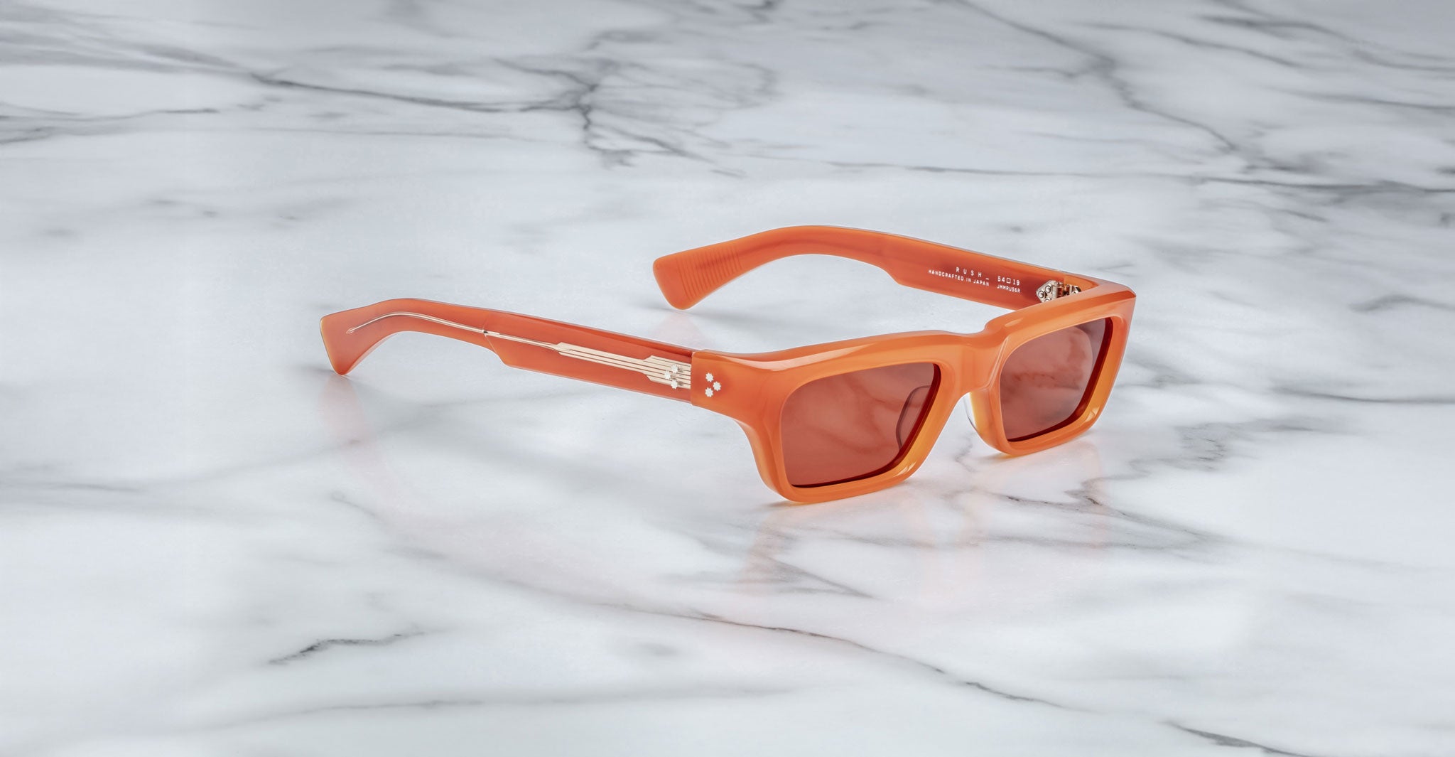 The Jacques Marie Mage Rush sunglasses, featuring an orange rectangular frame with tinted lenses, rest on a white marble surface. Metallic details and decorative elements accent the arms near the hinges.