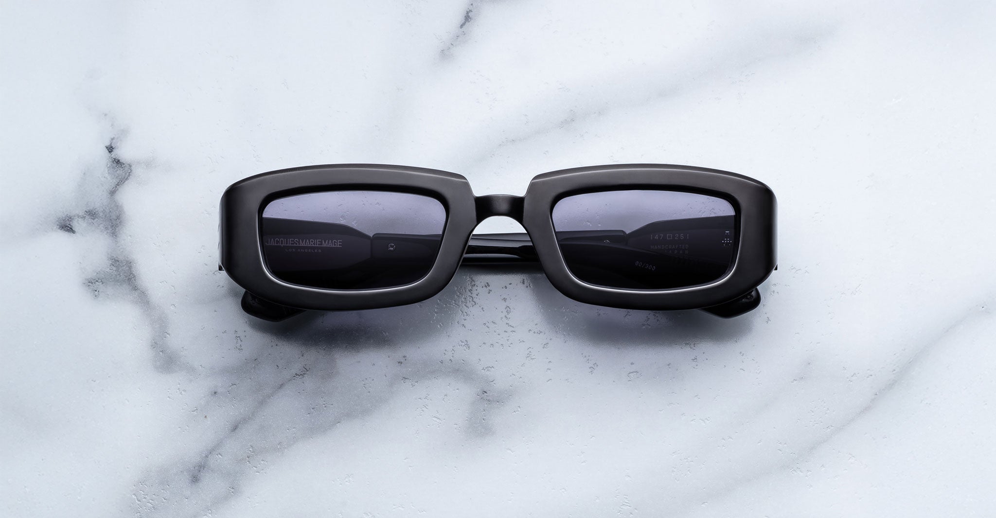 Jacques Marie Mage Ryder in Black, rectangular black acetate sunglasses with thick sculpted rims, a low-profile bridge, and dark tinted lenses, shown front-facing on a light marble surface.