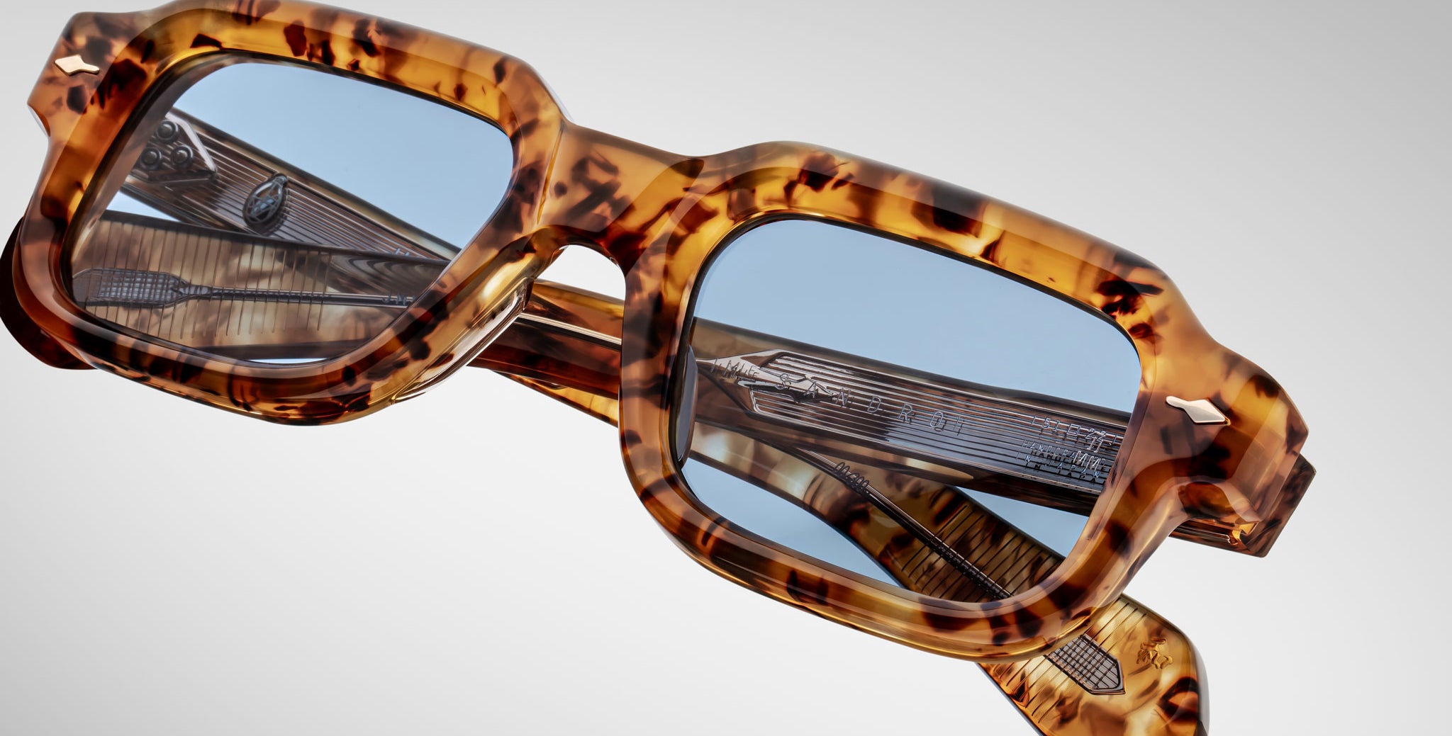 Close-up of Jacques Marie Mage Sandro eyeglasses with a thick, square tortoiseshell frame and blue-tinted lenses, resting against another pair with a translucent, textured frame.