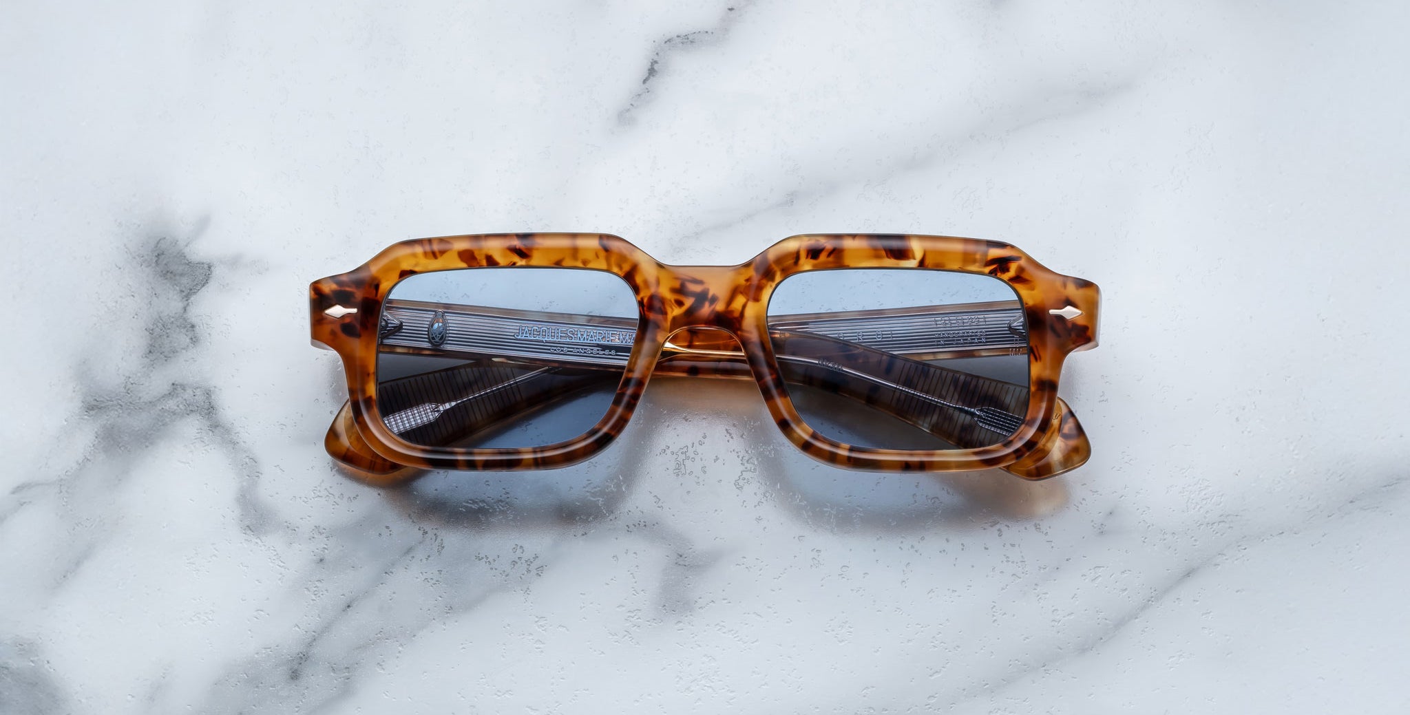 A pair of Jacques Marie Mage Sandro square tortoiseshell sunglasses with blue-tinted lenses rests on a white marble surface.