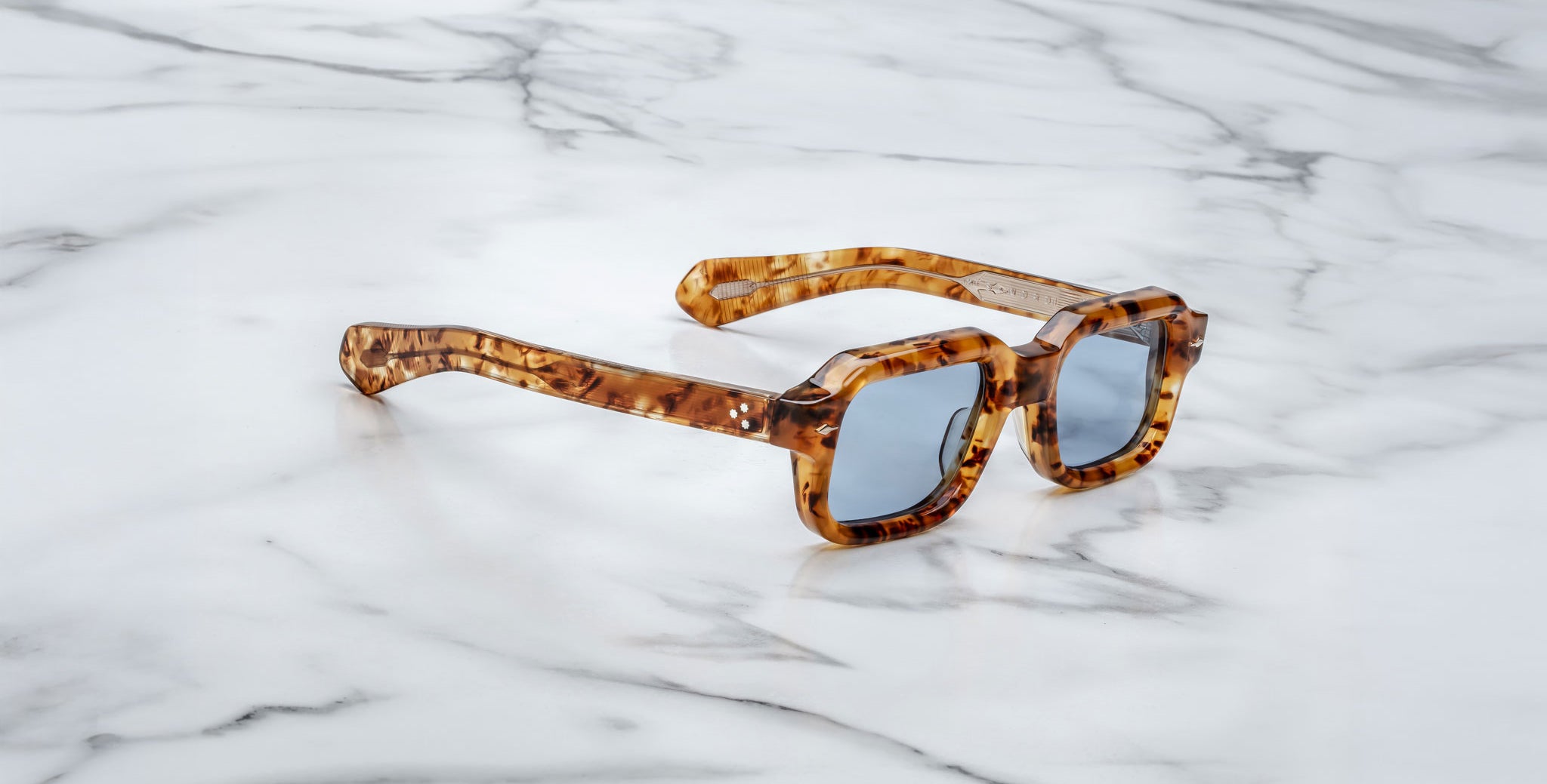 A pair of Jacques Marie Mage Sandro tortoiseshell square sunglasses with blue-tinted lenses rests on a white marble surface.