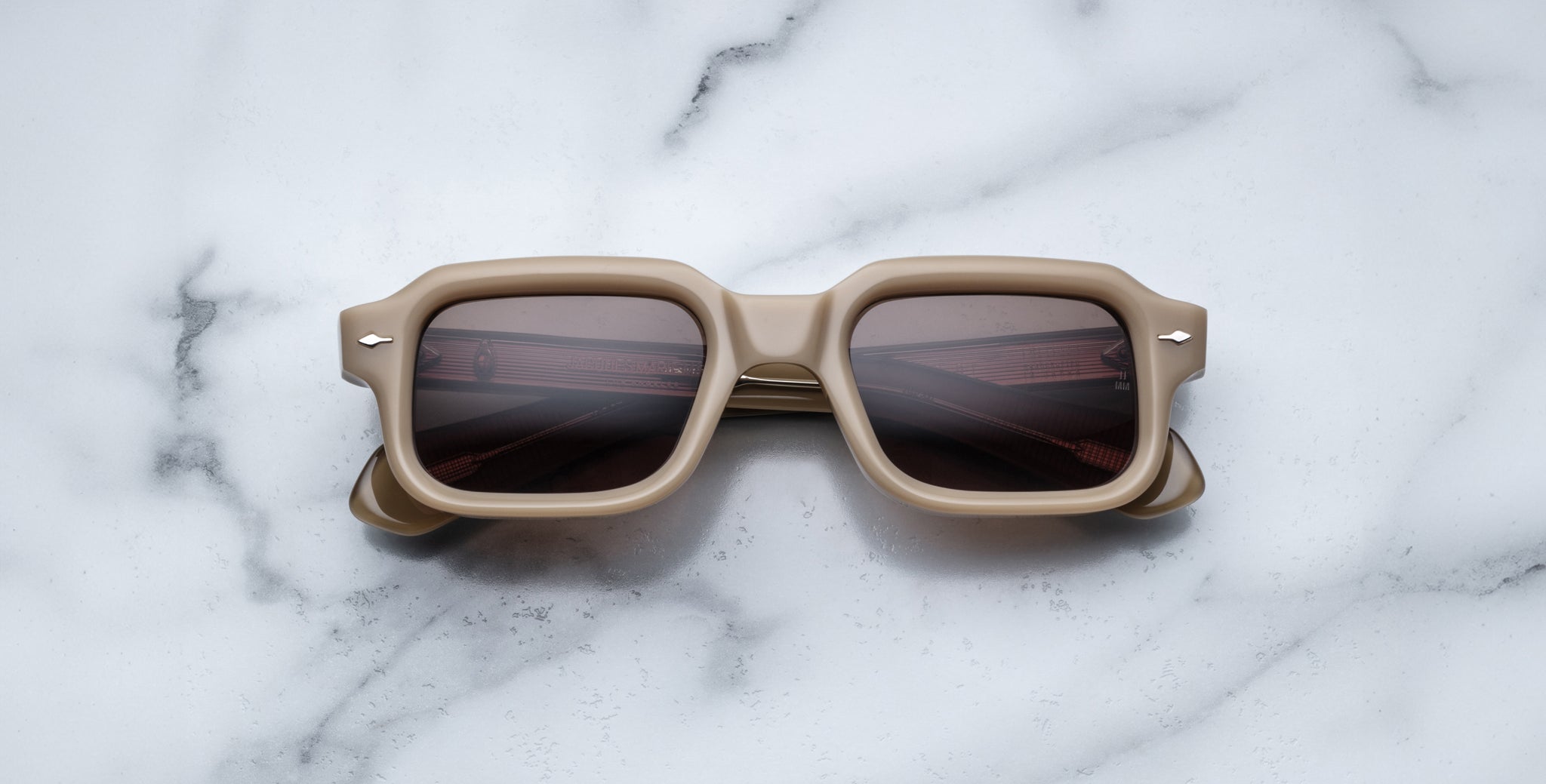 The Jacques Marie Mage Sandro sunglasses, featuring a square beige frame and dark lenses, rest on a white marble surface with gray veining.
