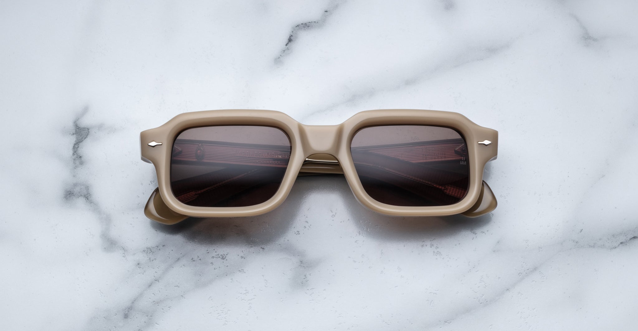 The Jacques Marie Mage Sandro sunglasses, featuring a square beige frame and dark lenses, rest on a white marble surface with gray veining.