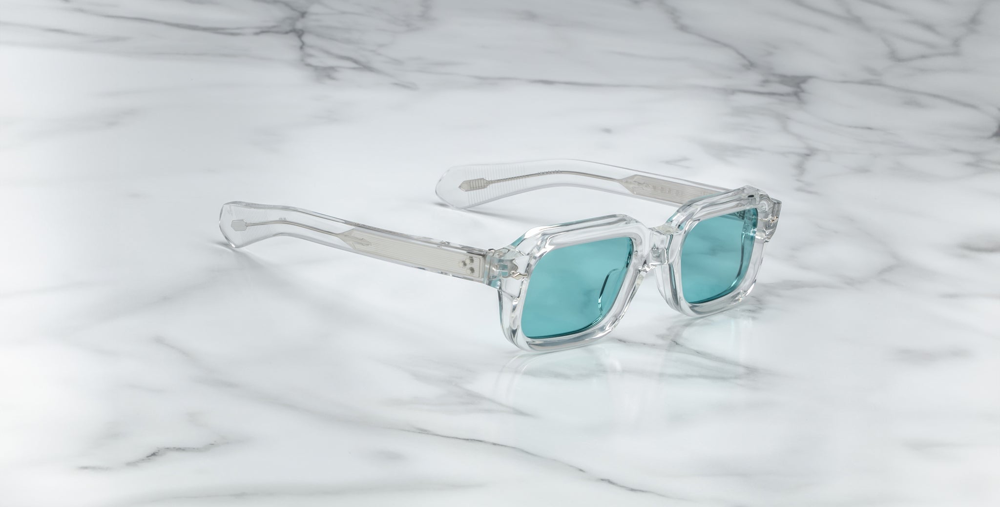 Jacques Marie Mage Sandro clear-framed sunglasses with teal lenses placed on a white marble surface featuring gray veining.