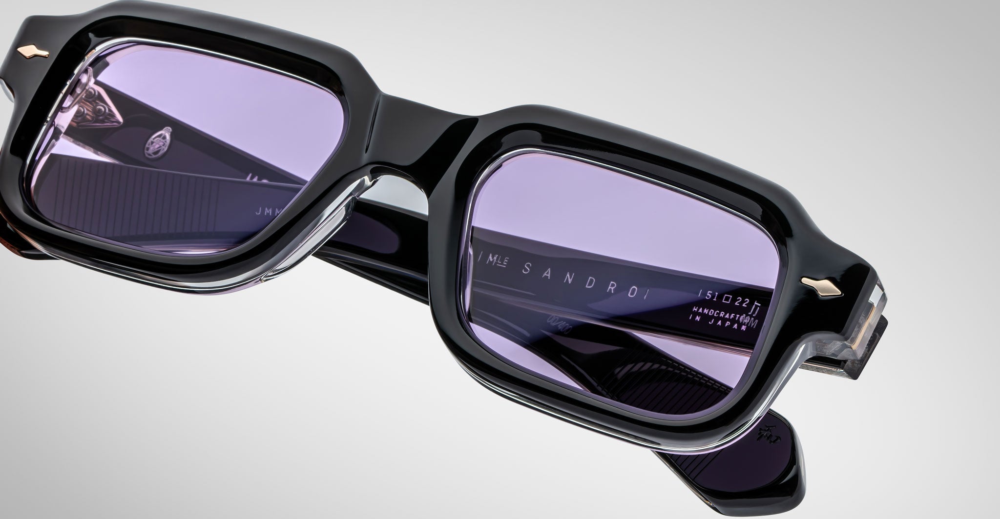 Close-up of Jacques Marie Mage Sandro black rectangular eyeglass frames with purple-tinted lenses. Sandro and Handmade in Japan are engraved inside the arms. Set against a plain gray background.