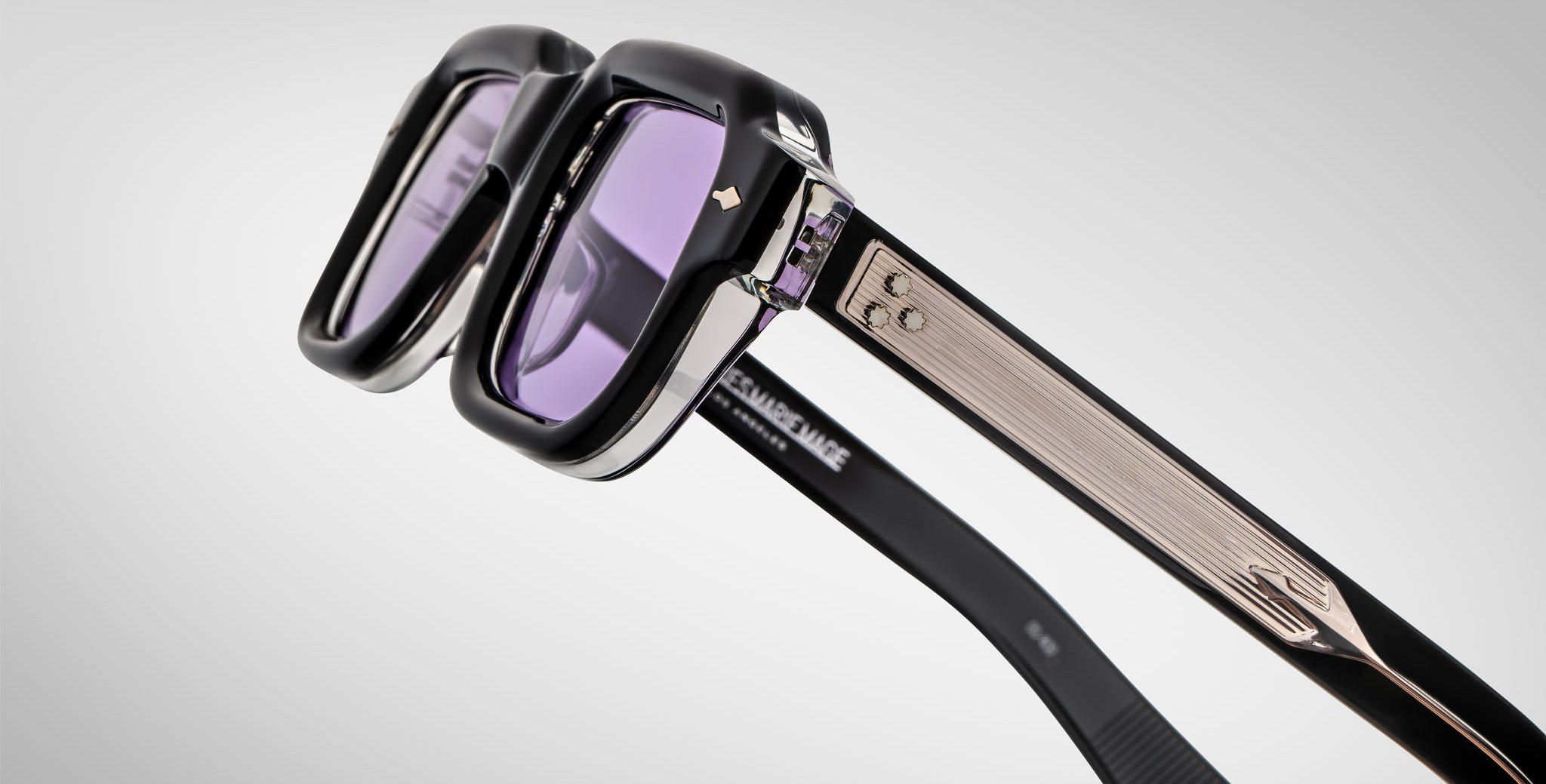 Close-up of Jacques Marie Mage Sandro sunglasses featuring thick black and silver frames, purple-tinted lenses, and textured metallic arms with decorative accents against a plain light background.