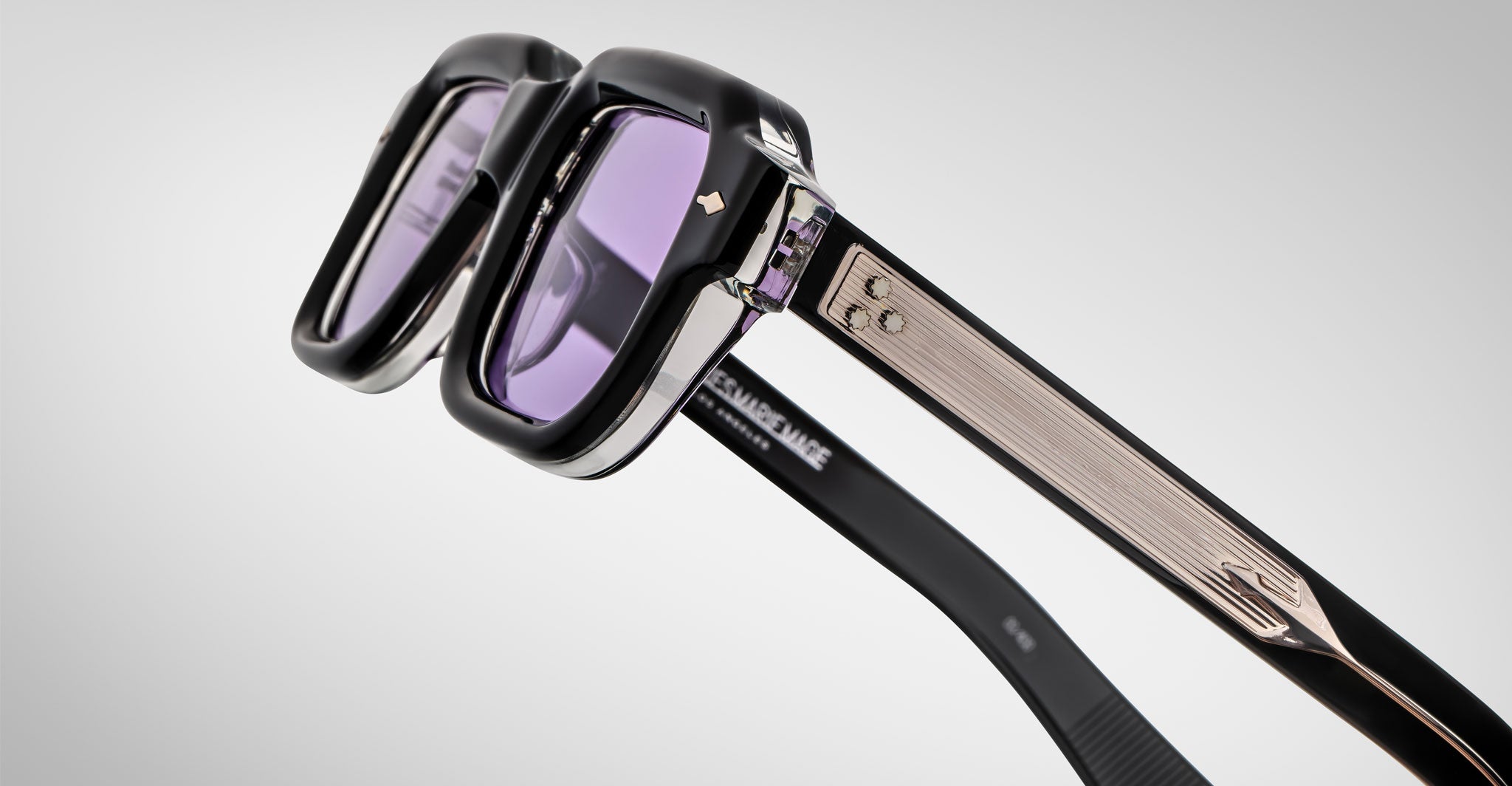 Close-up of Jacques Marie Mage Sandro sunglasses featuring thick black and silver frames, purple-tinted lenses, and textured metallic arms with decorative accents against a plain light background.