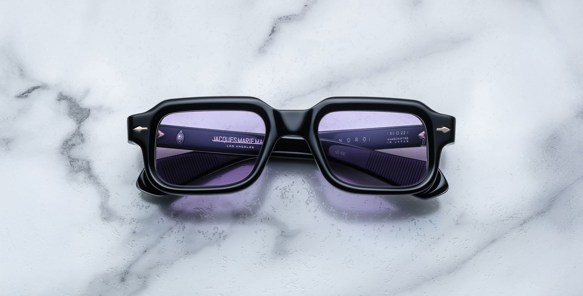 A pair of Jacques Marie Mage Sandro black rectangular sunglasses with purple-tinted lenses rests on a white and gray marble surface. Branding details are printed in small white text on the left lens.