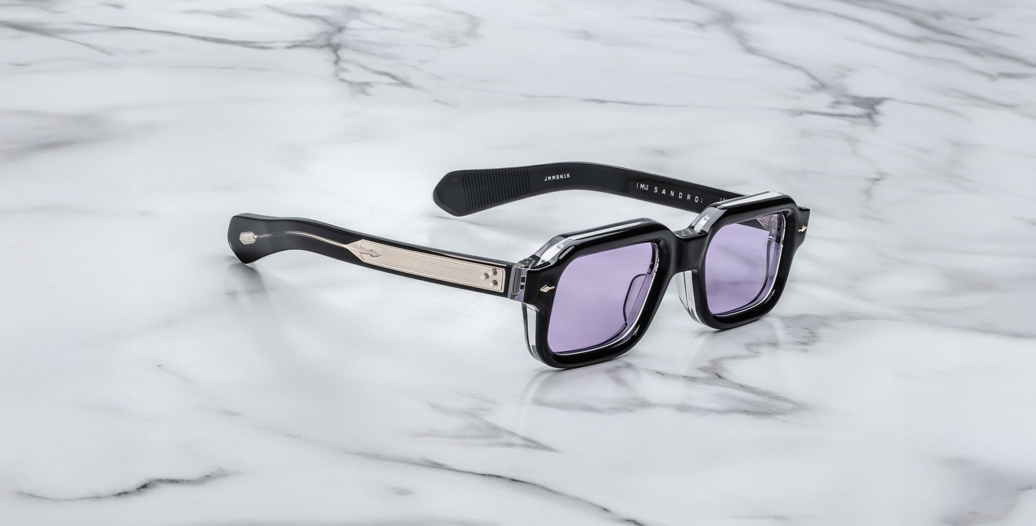 The Jacques Marie Mage Sandro sunglasses feature a black rectangular frame, light purple lenses, and metallic temple detailing, showcased on a white marble surface.