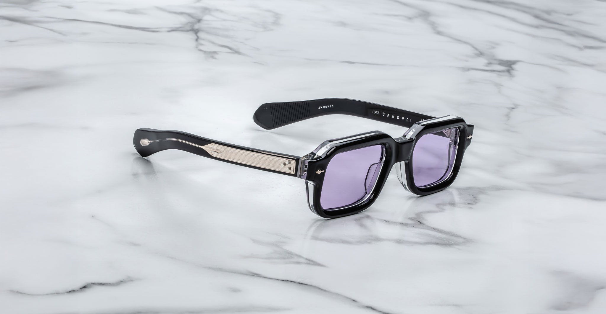 The Jacques Marie Mage Sandro sunglasses feature a black rectangular frame, light purple lenses, and metallic temple detailing, showcased on a white marble surface.