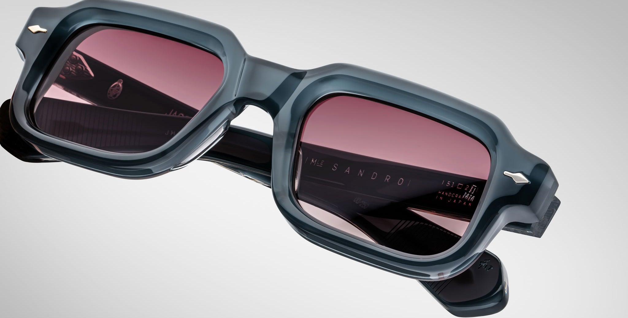 Close-up of Jacques Marie Mage Sandro sunglasses with thick, dark square frames and pink-tinted lenses, set against a light gray background. The brand name appears on the inside of one arm.