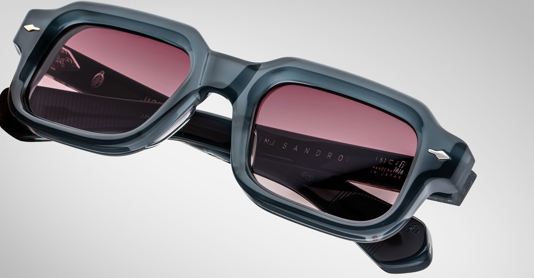 Close-up of Jacques Marie Mage Sandro sunglasses with thick, dark square frames and pink-tinted lenses, set against a light gray background. The brand name appears on the inside of one arm.