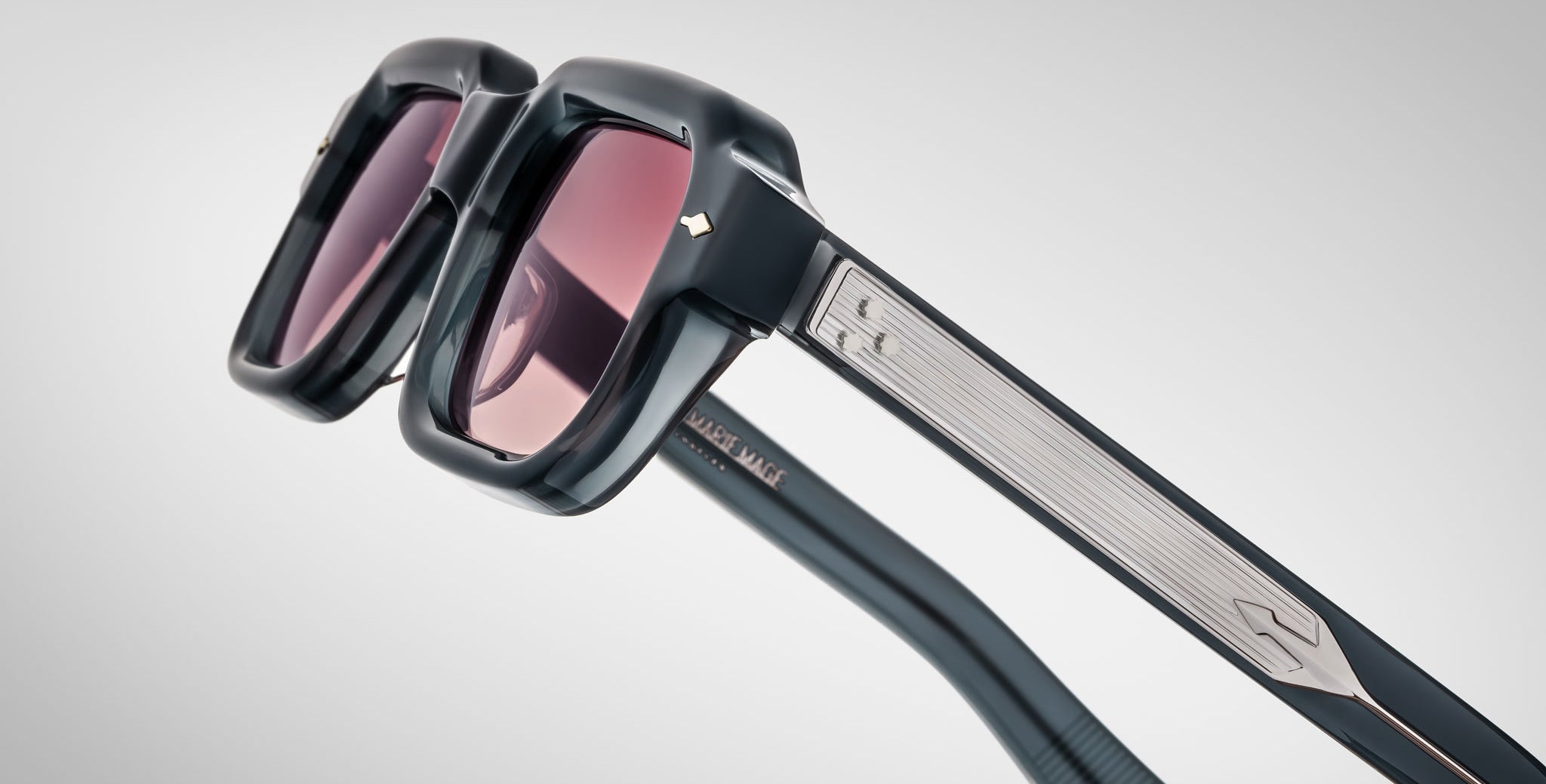 Close-up of Jacques Marie Mage Sandro sunglasses: black, thick frames with pink-tinted lenses. The arms display metallic accents, a textured pattern, and a small metallic logo near the hinge.