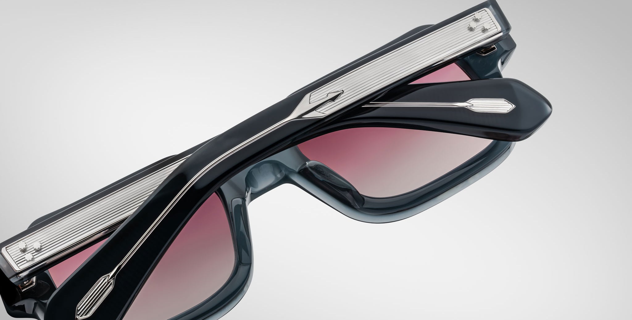 A close-up of Jacques Marie Mage Sandro sunglasses with thick, dark frames and pink-tinted lenses. The metal arms have a textured pattern and arrow-shaped detail near the hinges, set against a plain light gray background.