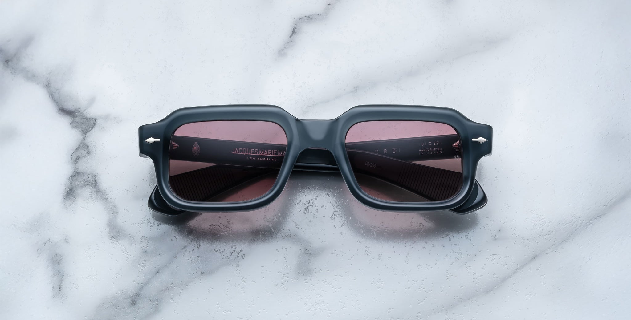 A pair of Jacques Marie Mage Sandro square sunglasses with black frames and dark red lenses rests on a white marble surface with grey veining.