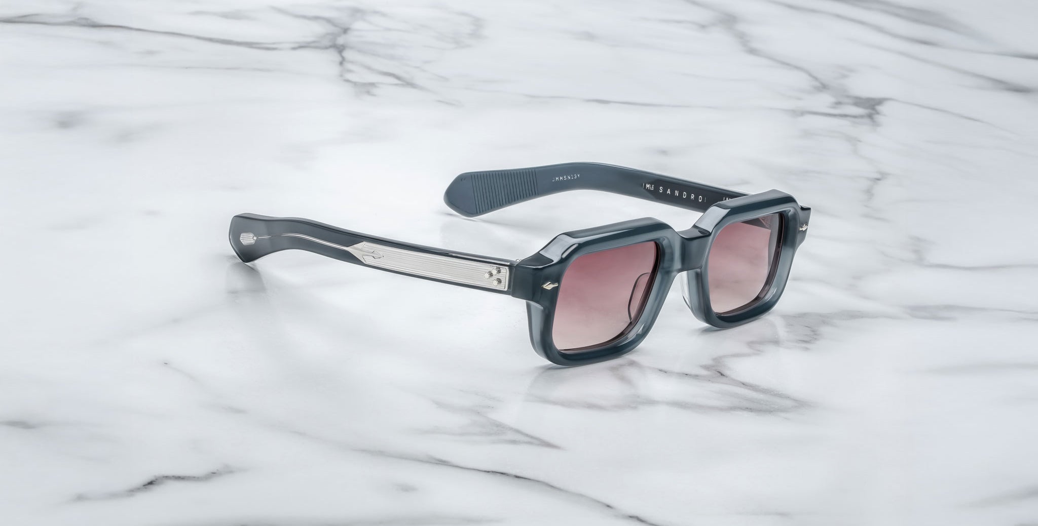 A pair of Jacques Marie Mage Sandro sunglasses with black square frames, metallic arm details, and pink-tinted lenses rests on a white marble surface.